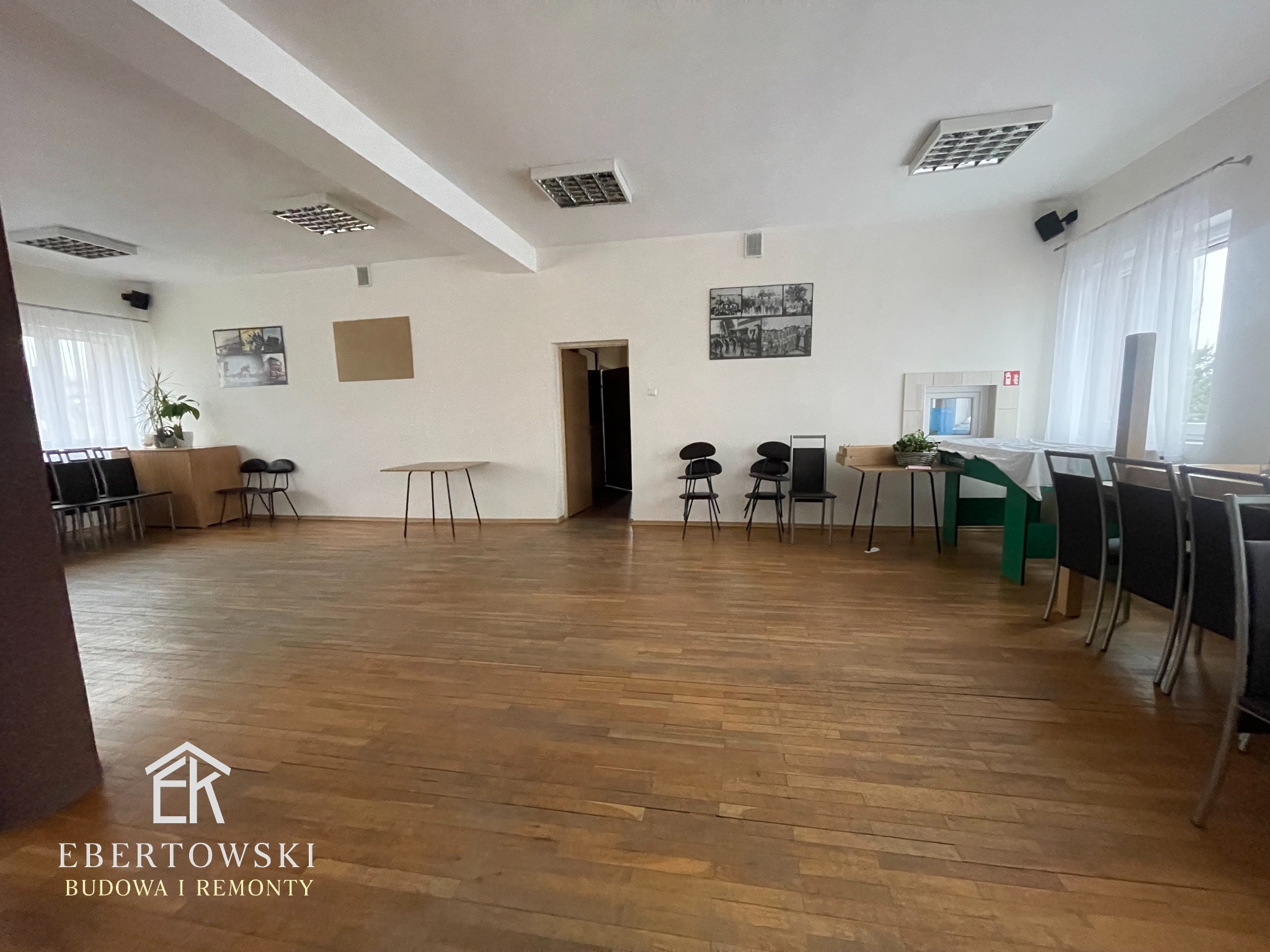 Sala po remoncie z drewnianą podłogą, białymi ścianami i sufitami. Widoczne krzesła, stoły i oświetlenie. Na ścianach wiszą obrazy. Charakterystyczny wzór parkietu.