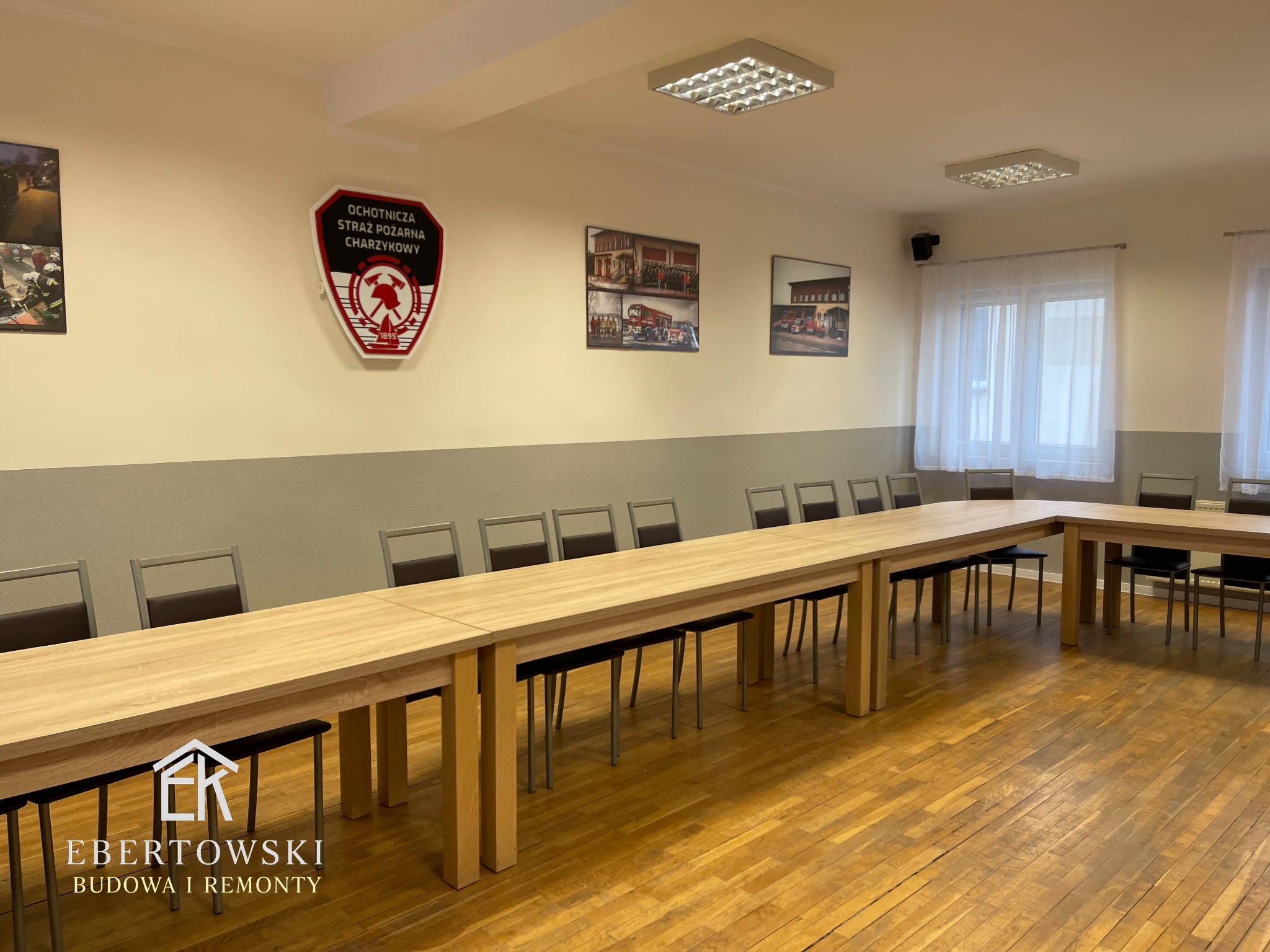 Jasne, odnowione wnętrze sali konferencyjnej z długim stołem i krzesłami. Na ścianie logo OSP i zdjęcia. Parkiet po renowacji. Widoczny logotyp firmy remontowej.