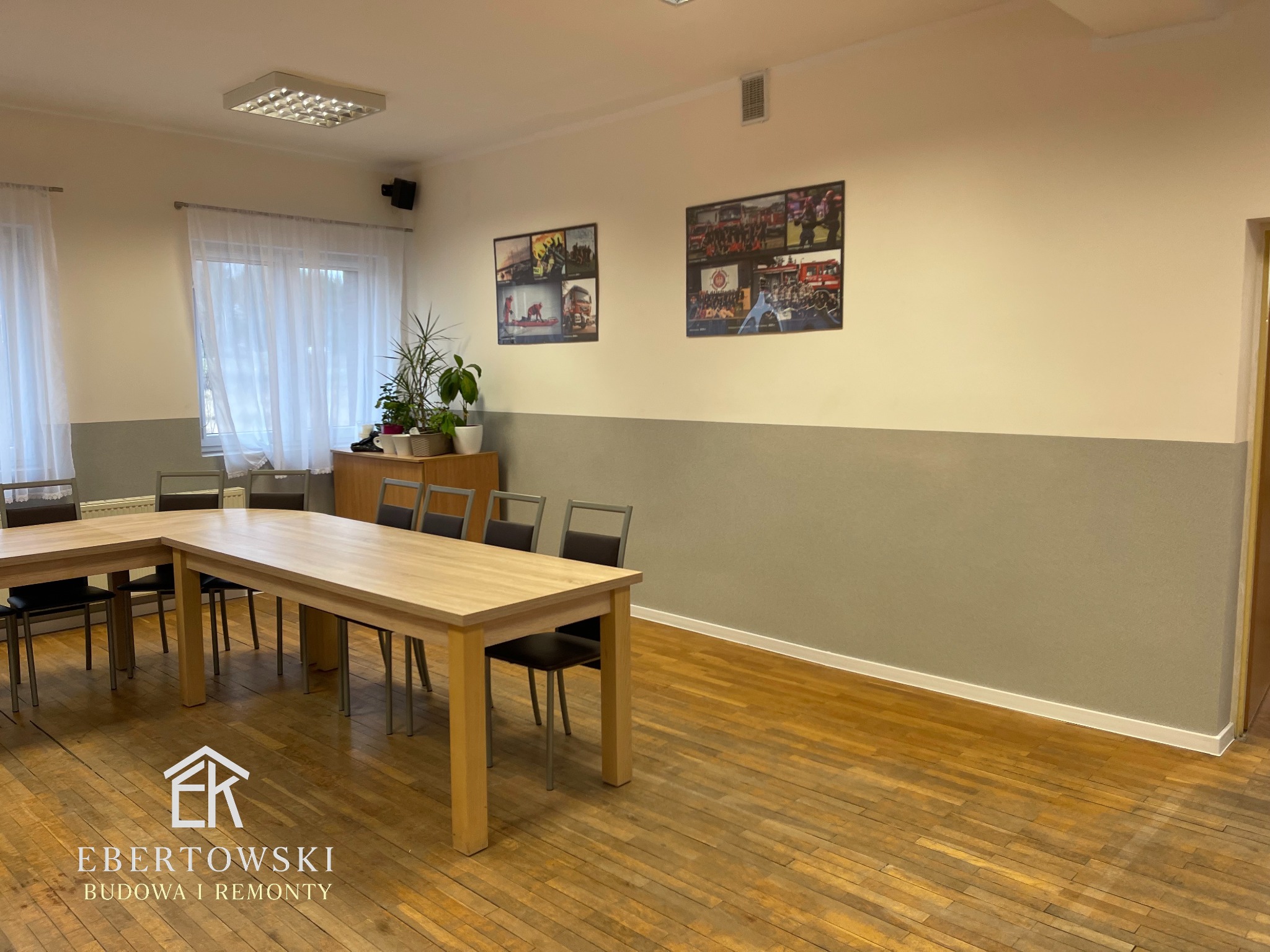 Jasne biuro po odświeżeniu: drewniana podłoga, stół konferencyjny, szare ściany do połowy wysokości, zdjęcia w ramkach na górze. Widok na okno z białymi firankami.