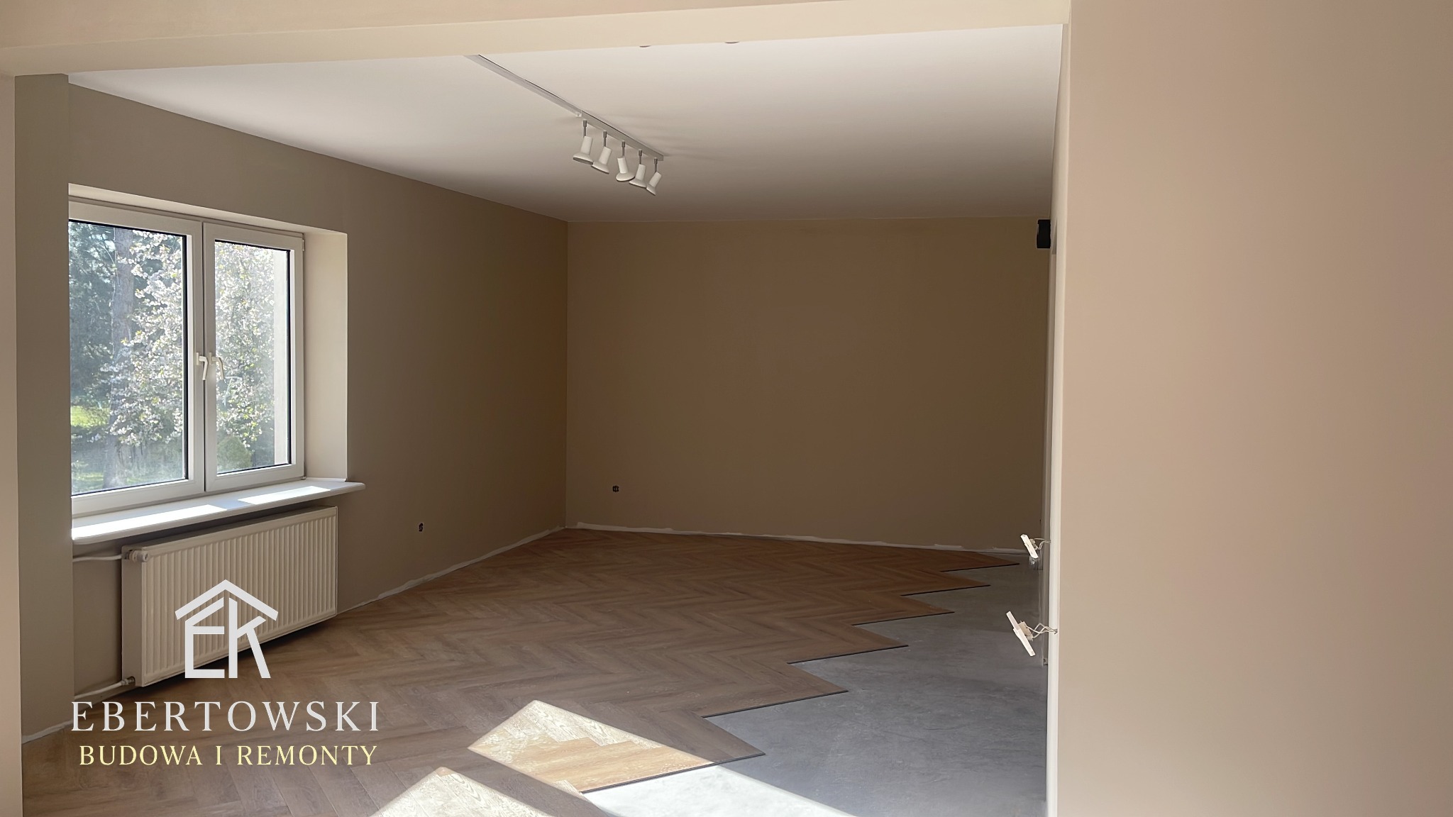 Układanie parkietu w jodełkę w pokoju z beżowymi ścianami i białym oknem. Część podłogi jeszcze nieukończona, widoczny surowy beton. Na ścianie widoczne oświetlenie.