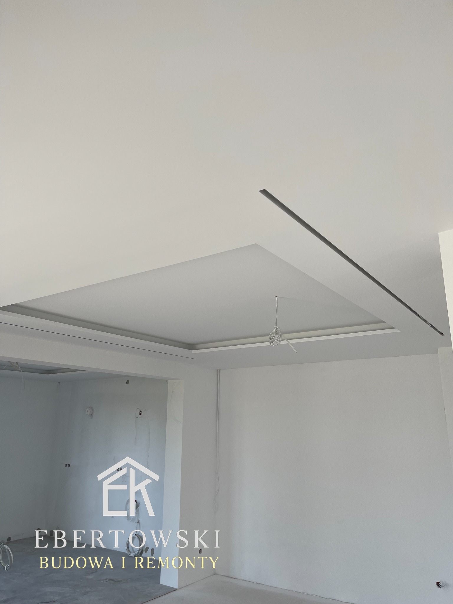 Biały sufit podwieszany z oświetleniem liniowym w nowym wnętrzu. Widoczne kable i logo firmy remontowej w dolnej części kadru. Minimalistyczny design.