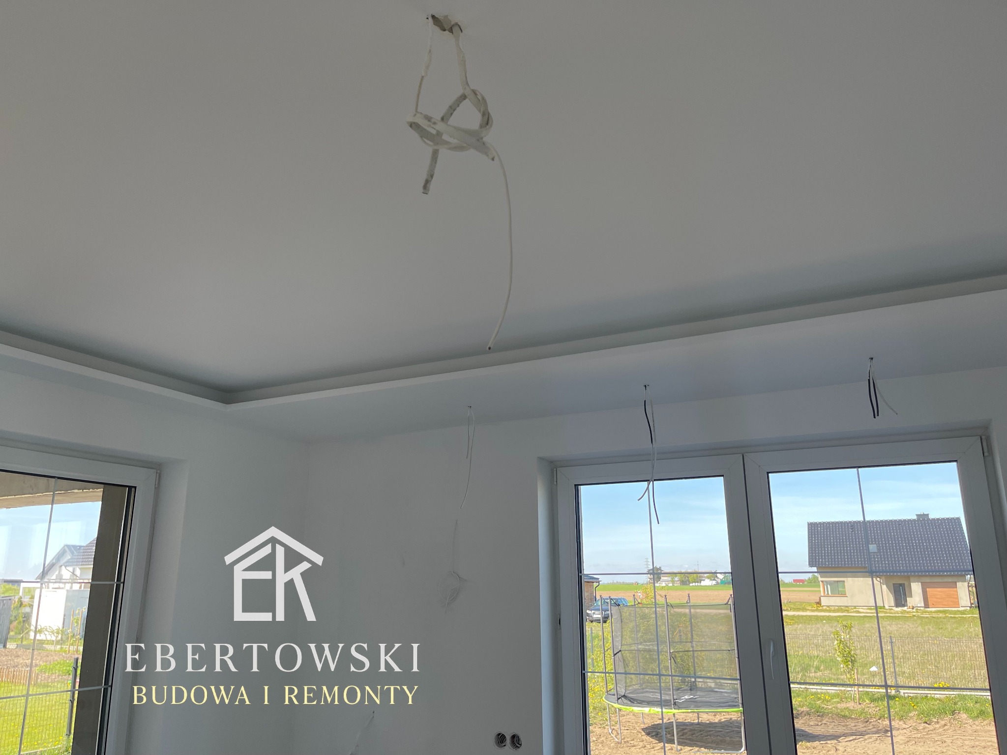 Białe wnętrze po remoncie z widocznymi przewodami elektrycznymi, listwami sufitowymi i oknami. Na ścianie logo firmy budowlanej.
