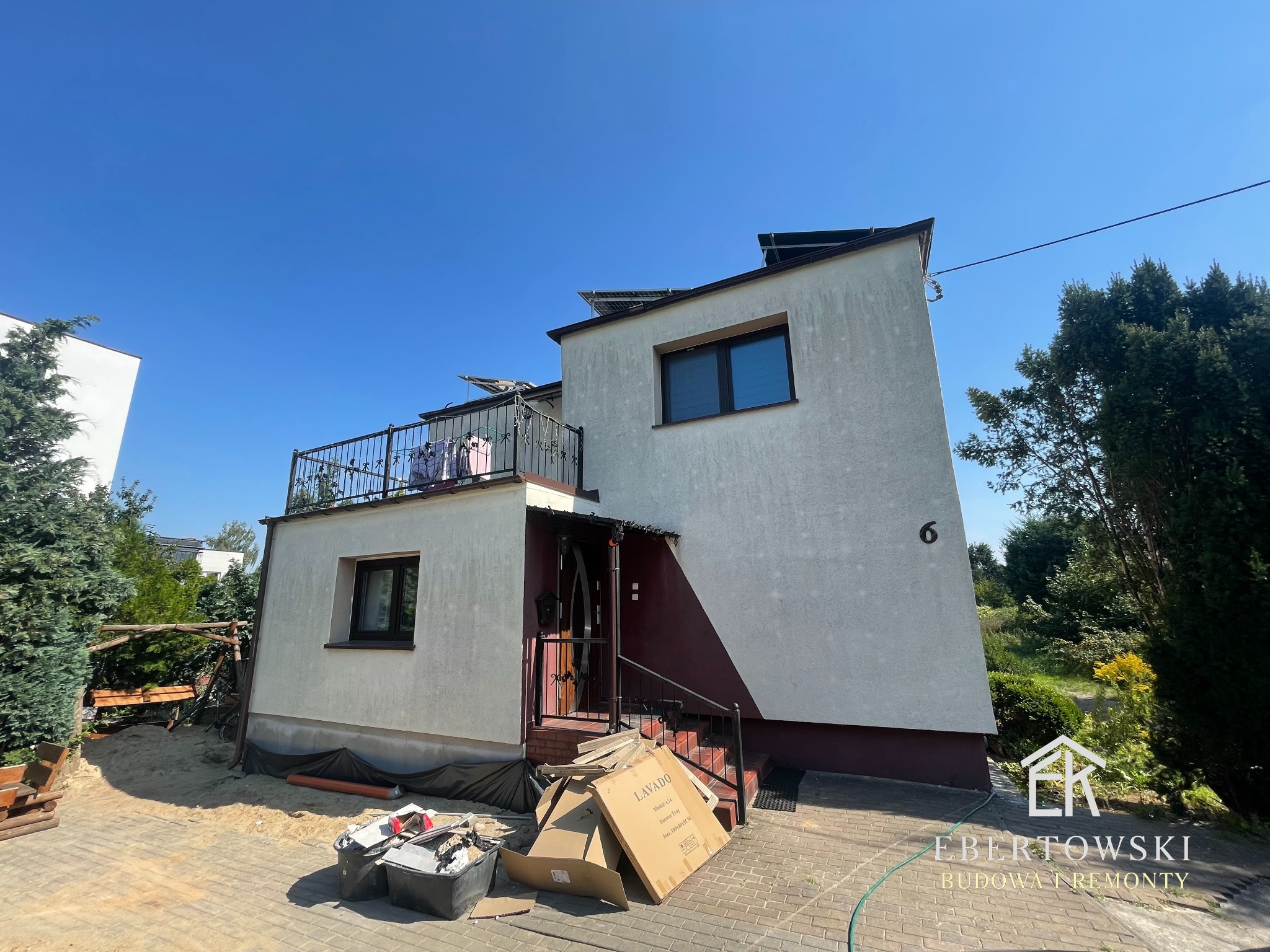 Dwupiętrowy dom z balkonem i panelami słonecznymi, widoczny z poziomu chodnika. Na pierwszym planie kartony i pojemniki, sugerujące prace remontowe.
