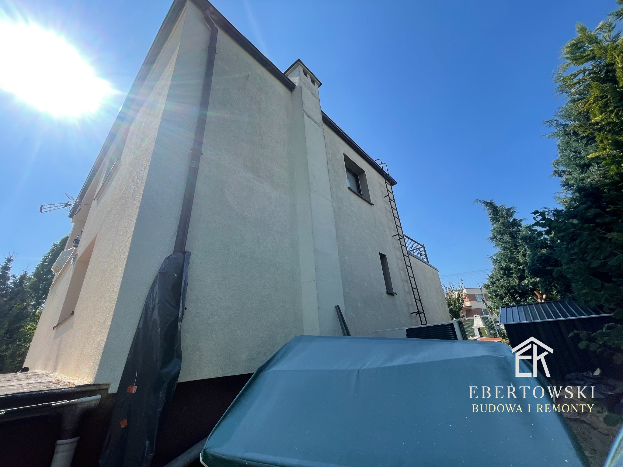 Elewacja domu jednorodzinnego po remoncie, widok z dołu, błękitne niebo, fragment dachu garażu z logo firmy budowlanej w prawym dolnym rogu.