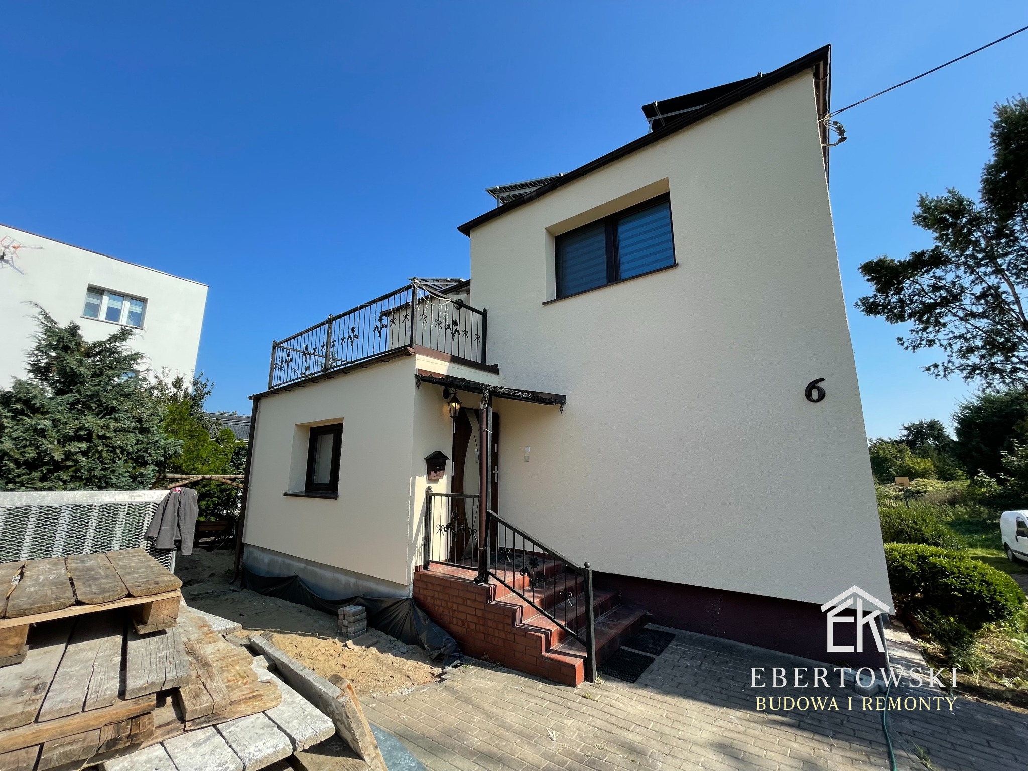 Elewacja domu jednorodzinnego w jasnym odcieniu z balkonem i schodami wejściowymi. Widoczne palety i materiały budowlane. Błękitne niebo w tle.