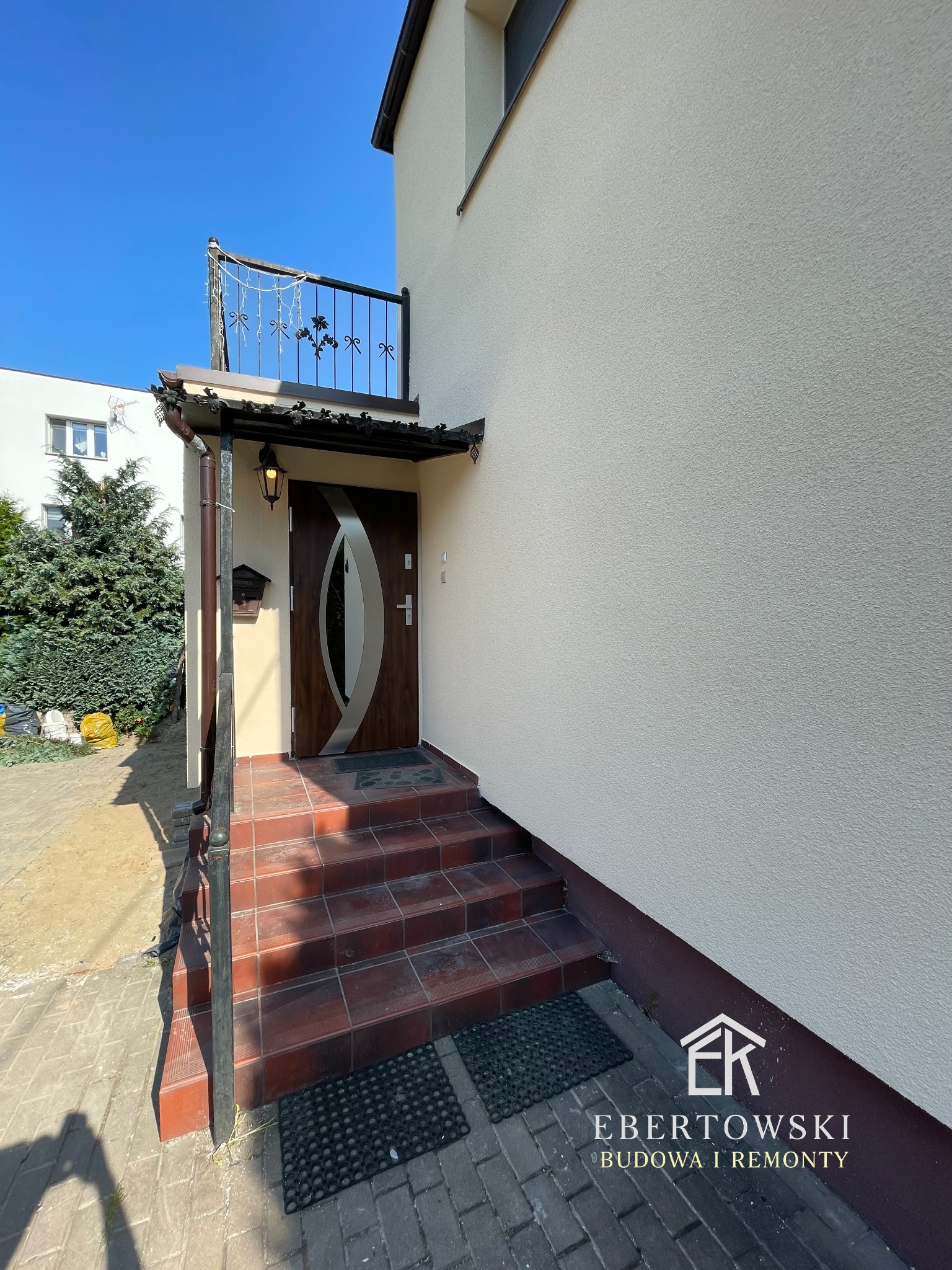 Elewacja domu po remoncie: jasna ściana z wejściem, brązowe drzwi z owalnym przeszkleniem, schody z terakoty i balustrada. Widoczny fragment balkonu z ozdobną barierką.