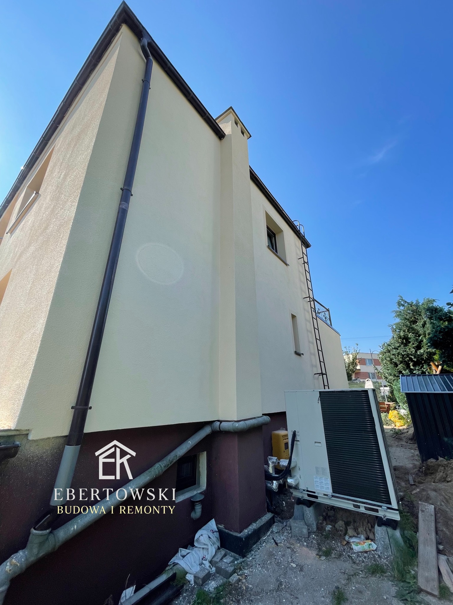 Elewacja domu po remoncie, z widoczną nową, jasną kolorystyką i detalami wykończeniowymi. Widoczny fragment instalacji zewnętrznej i logo firmy.
