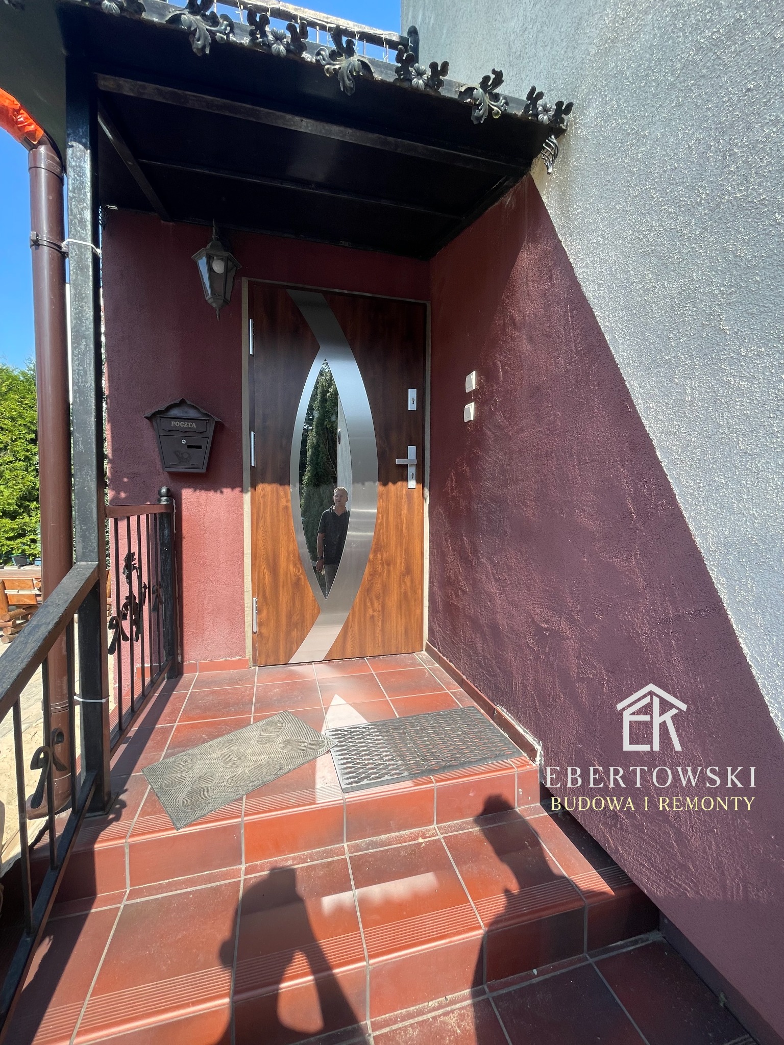 Wejście do domu z nowoczesnymi drzwiami z owalnym przeszkleniem, brązową elewacją i terakotą na schodach. Widoczna skrzynka pocztowa i cień osoby.