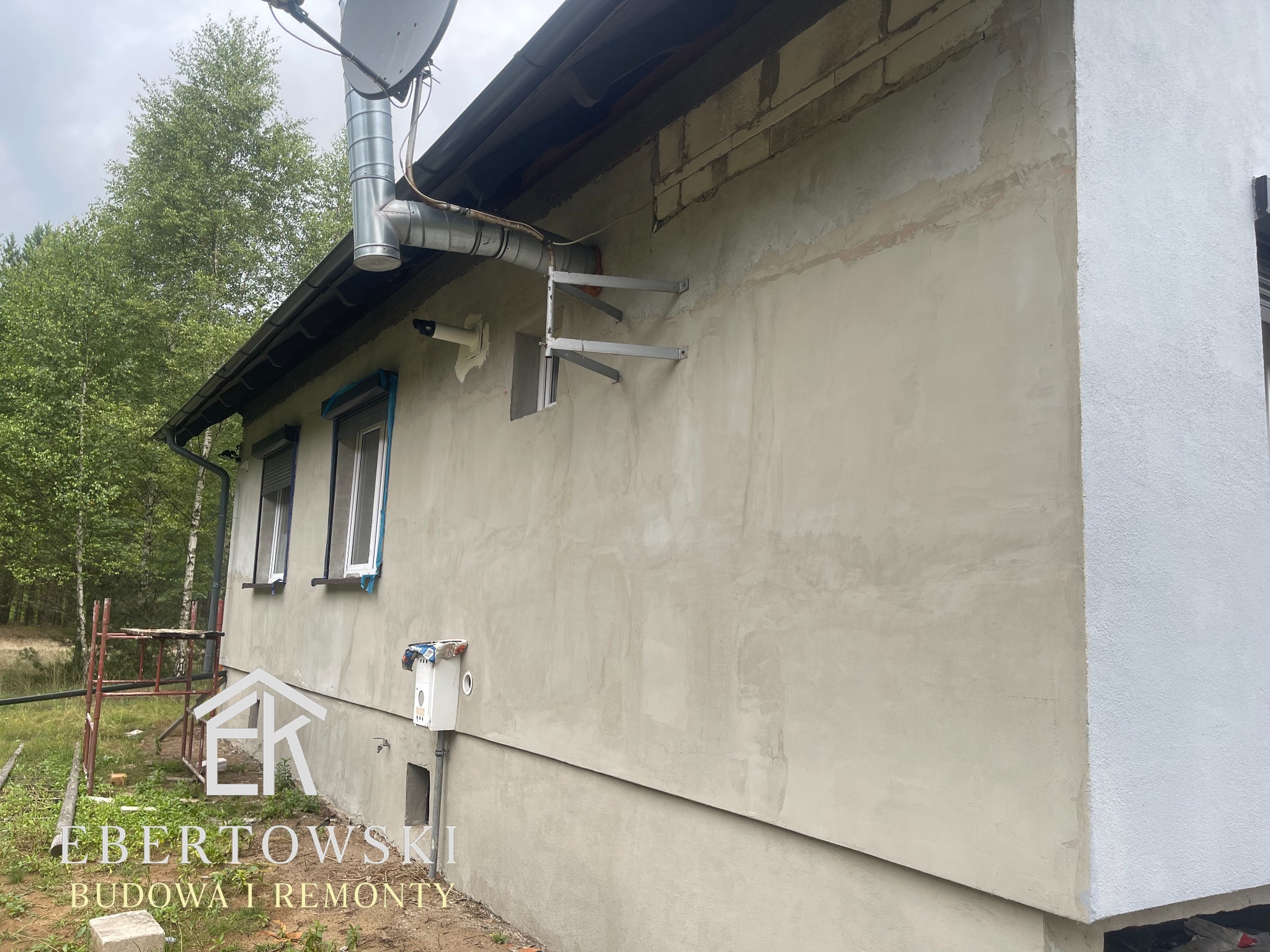 Ściana domu w trakcie prac wykończeniowych: fragment otynkowany, fragment pomalowany na biało. Widoczne rusztowanie i elementy instalacji na elewacji.