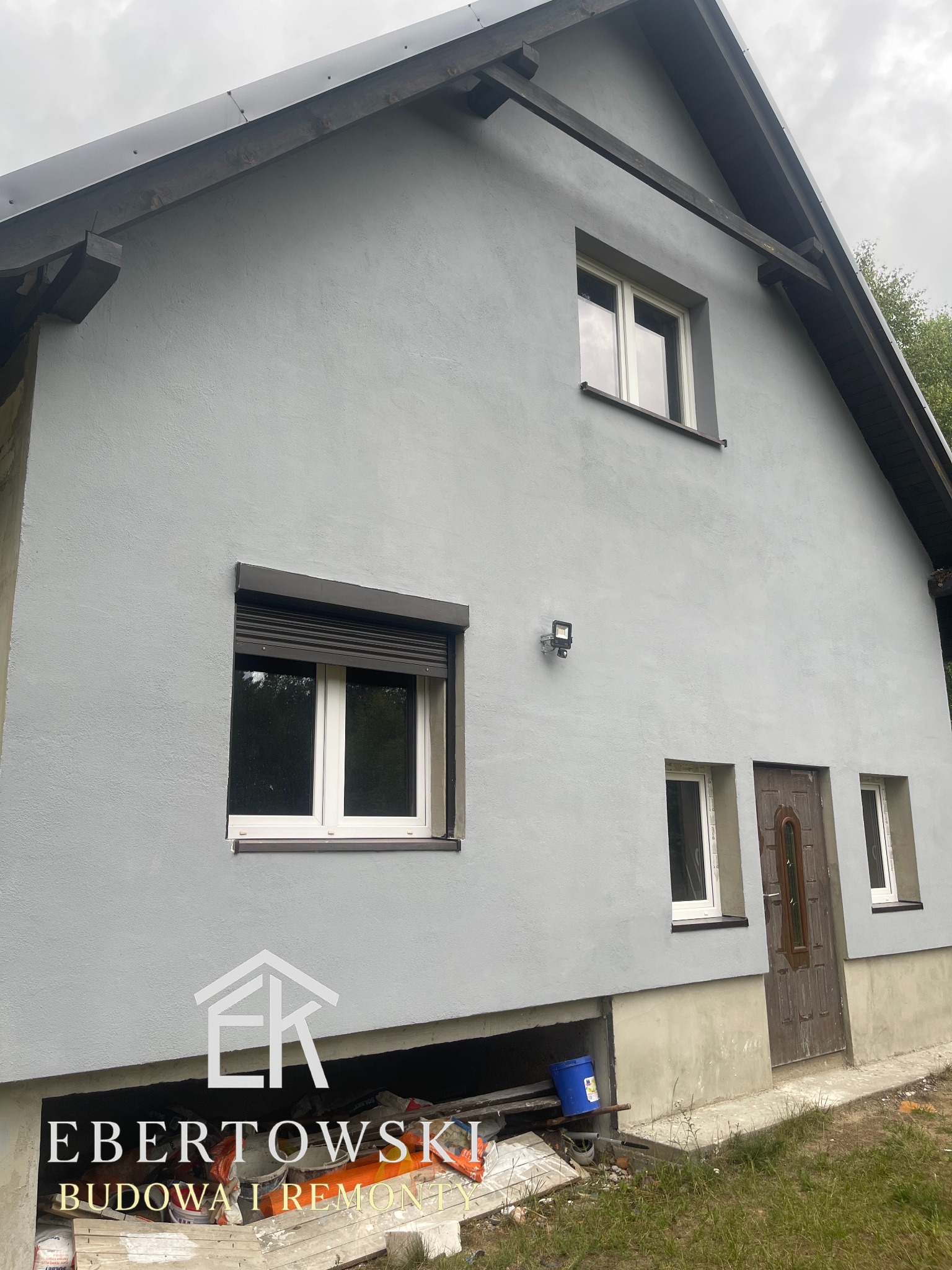 Elewacja domu po remoncie: szara ściana z oknami (jedno z roletą), drewniane elementy dachu. Widoczny fragment fundamentu z materiałami budowlanymi.