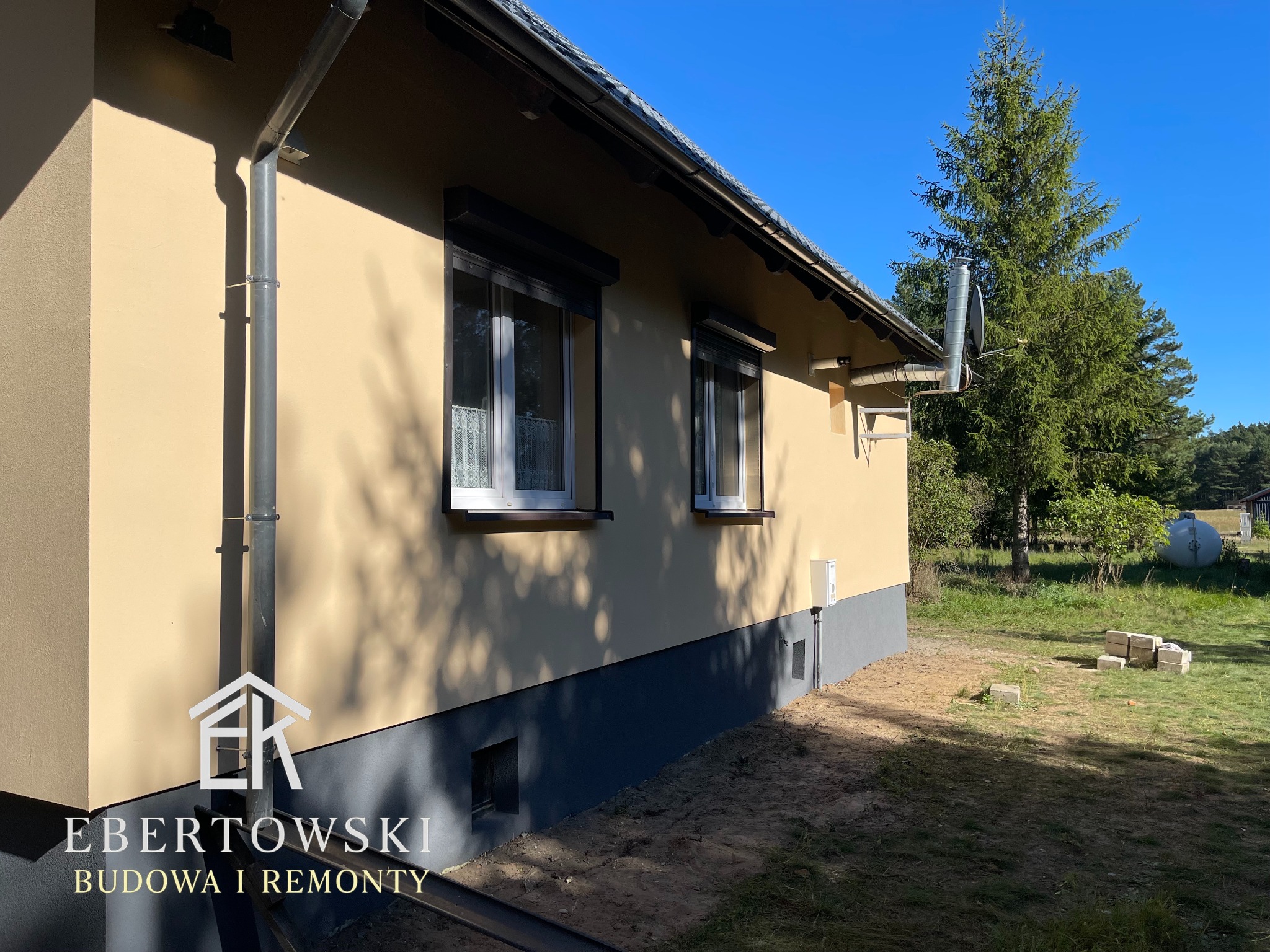 Ocieplony dom jednorodzinny z beżową elewacją, szarą podmurówką i ciemnymi roletami zewnętrznymi. Widoczny system rynnowy i komin. W tle drzewa i błękitne niebo.
