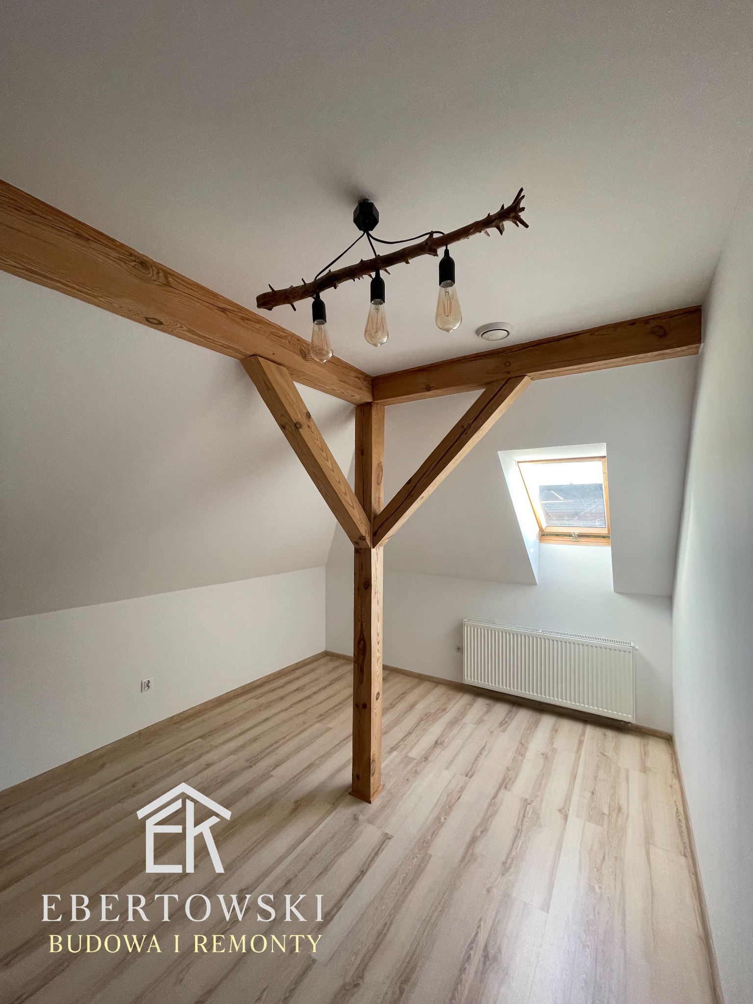 Jasne poddasze po remoncie z drewnianą konstrukcją nośną, oknem dachowym i oryginalną lampą z gałęzi. Widoczny grzejnik i podłoga imitująca drewno.