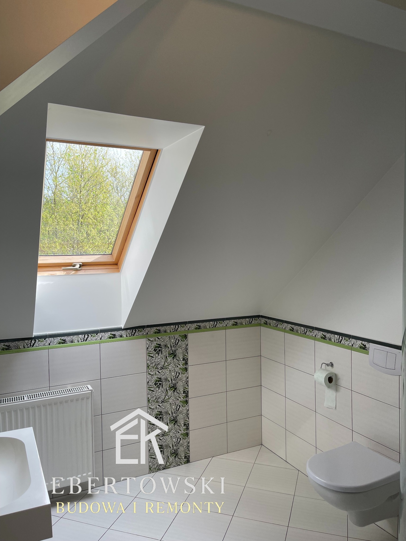 Jasna łazienka na poddaszu z oknem dachowym, wykończona białymi płytkami z zielonymi akcentami. Widoczny fragment umywalki, grzejnik i toaleta. Ujęcie z dołu.