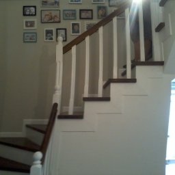 Stolpod - Wnętrze z drewnianymi schodami z białą balustradą, na ścianie galeria ramek ze zdjęciami, widoczne nogi osoby wchodzącej po schodach.