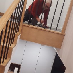 Stolpod - Montaż balustrady z czarnymi, pionowymi prętami na drewnianych schodach przez mężczyznę w czerwonej bluzie, widok z góry na szafę wnękową pod schodami.
