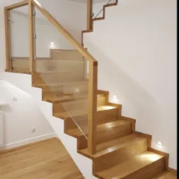 Jasne, drewniane schody z przeszkloną balustradą i punktowym oświetleniem na ścianie, z widocznym fragmentem podłogi wyłożonej parkietem.