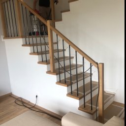 Stolpod - Montaż schodów z drewnianymi stopniami i metalowymi balustradami. Widoczny pracownik z odkurzaczem na schodach. Jasne wnętrze, minimalistyczny design.