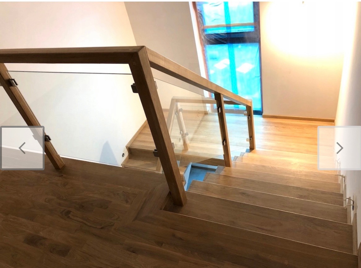 Nowoczesne schody z drewnianymi stopniami i balustradą ze szkła hartowanego z drewnianą ramą, widok z góry na dół, okno w tle zasłonięte folią ochronną.