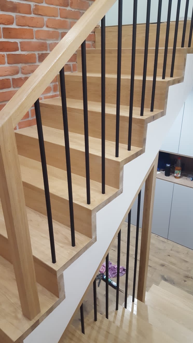 Jasne, drewniane schody z czarnymi, pionowymi balustradami, widok z góry, ściana z czerwonej cegły w tle.