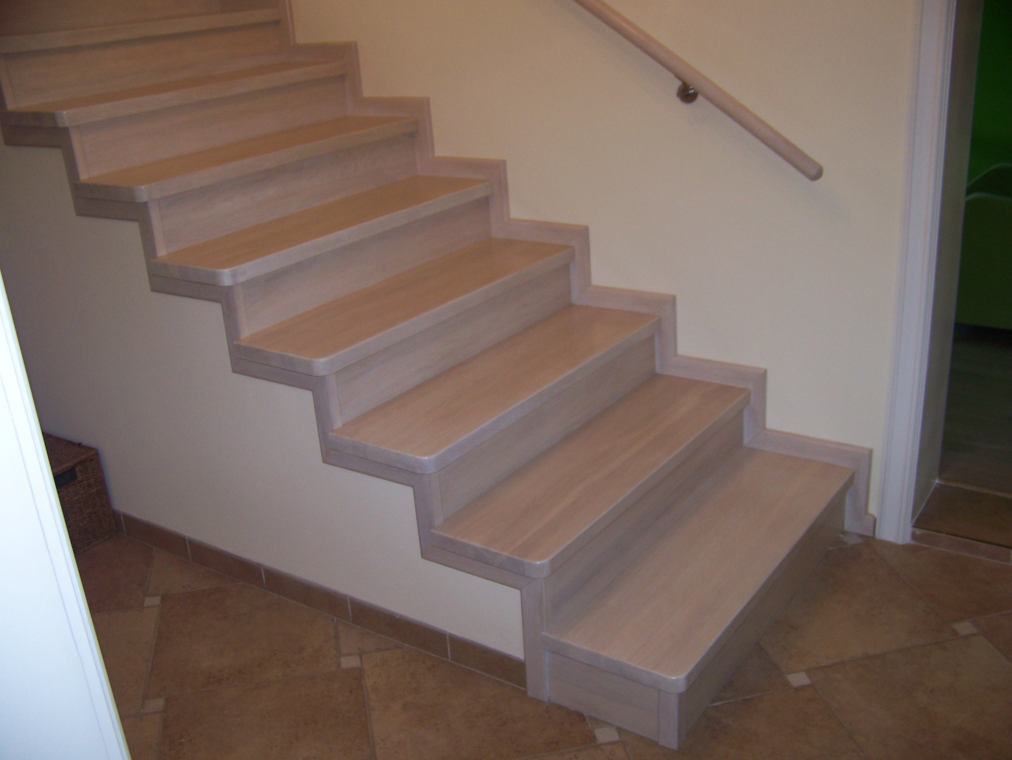 Jasne, drewniane schody z poręczą, widok z dołu na tle beżowej ściany i brązowej podłogi z płytek.