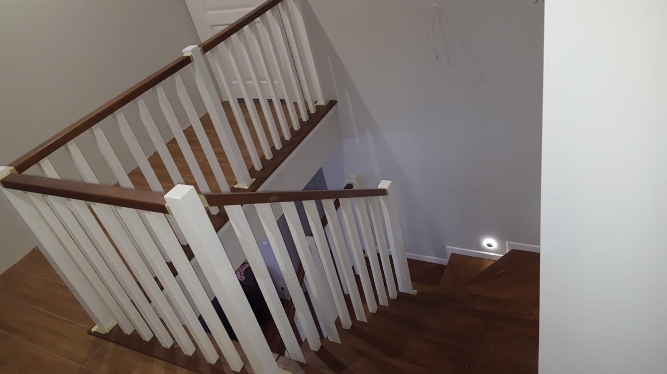 Wnętrze domu z drewnianymi schodami z białą balustradą i brązowymi poręczami, widoczne taśmy malarskie zabezpieczające elementy, oświetlenie punktowe w ścianie.