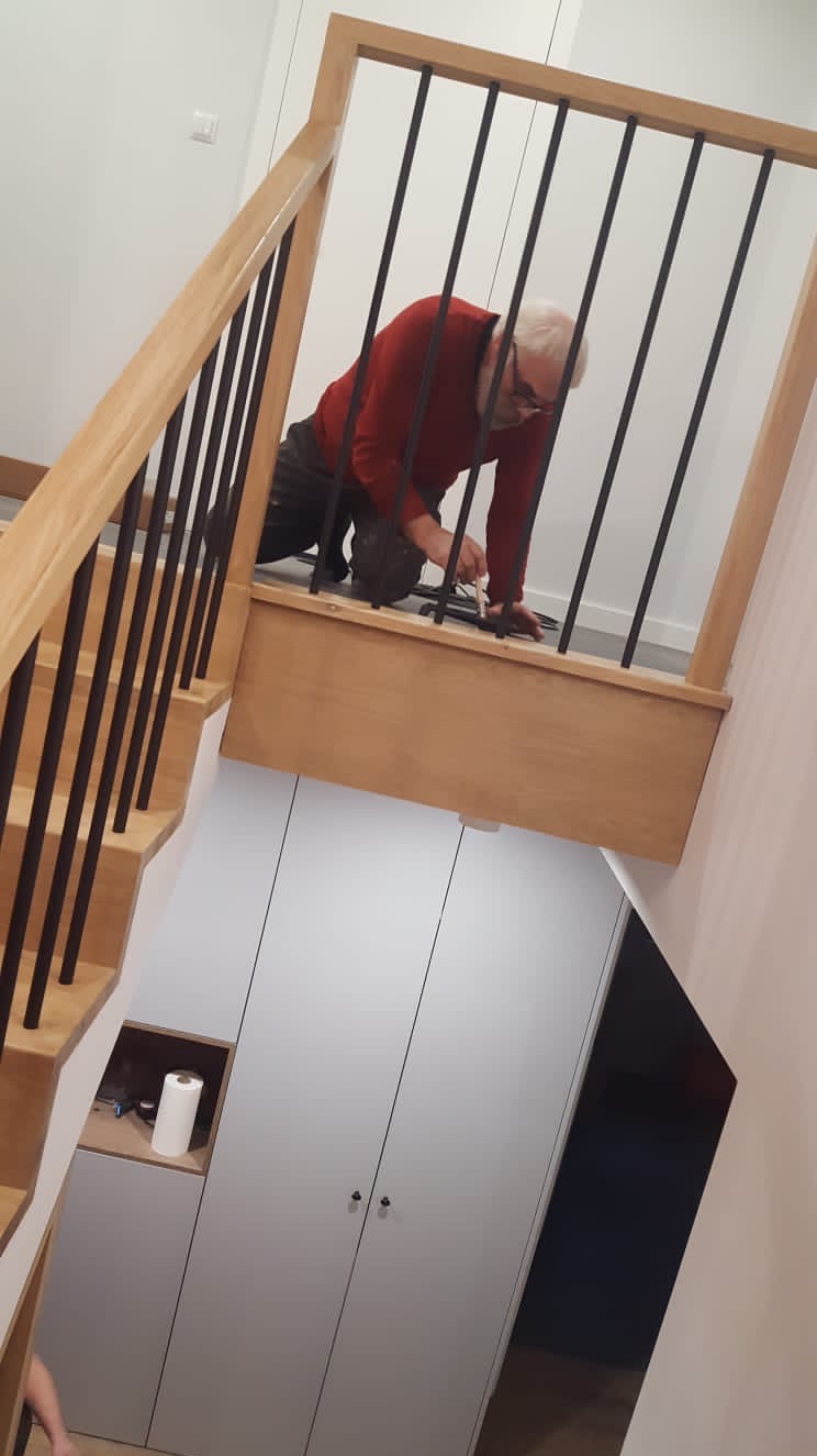 Montaż balustrady z czarnymi, pionowymi prętami na drewnianych schodach przez mężczyznę w czerwonej bluzie, widok z góry na szafę wnękową pod schodami.