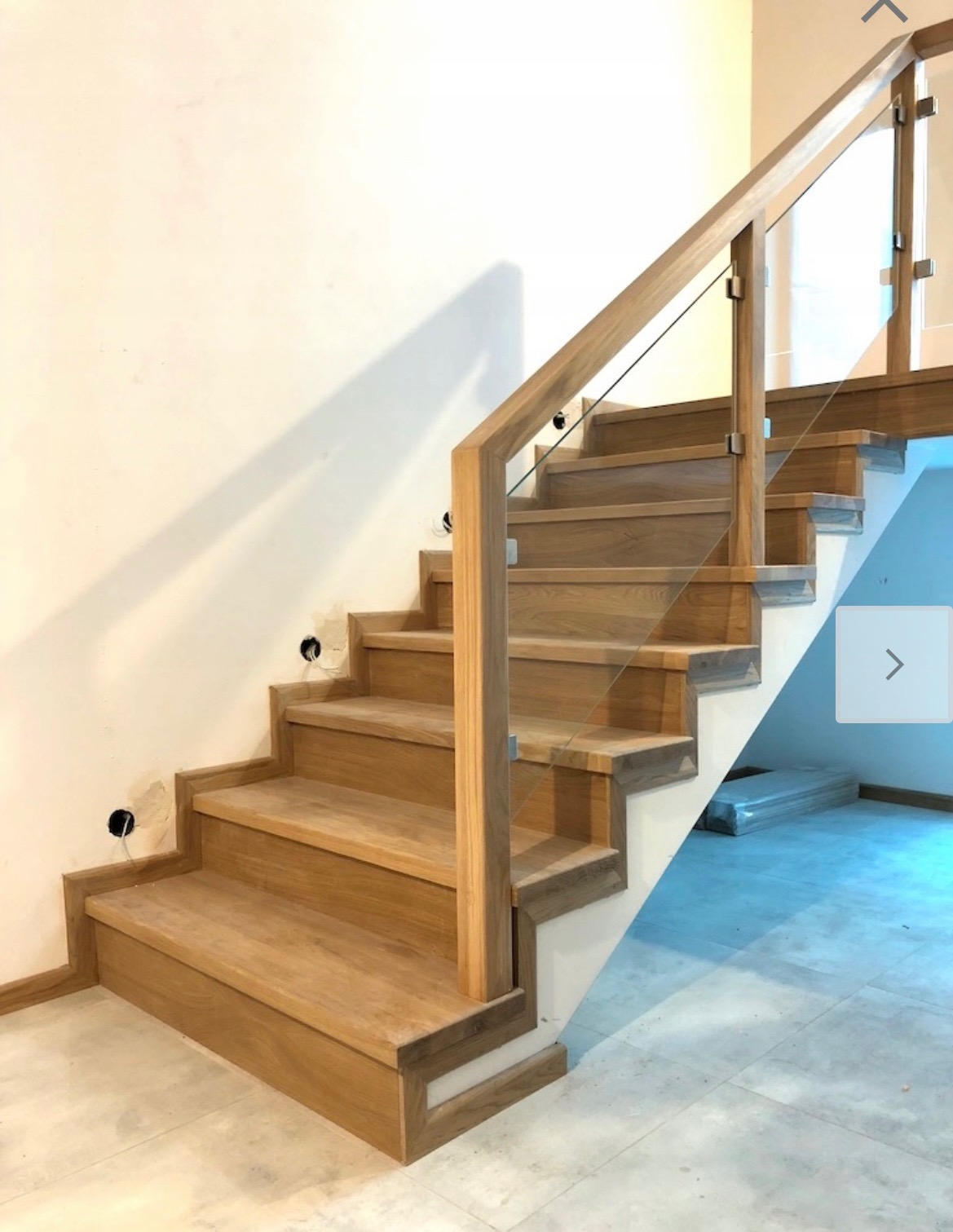 Jasne drewniane schody z balustradą ze szkła hartowanego, widoczne otwory pod gniazdka elektryczne w ścianie, minimalistyczny design.