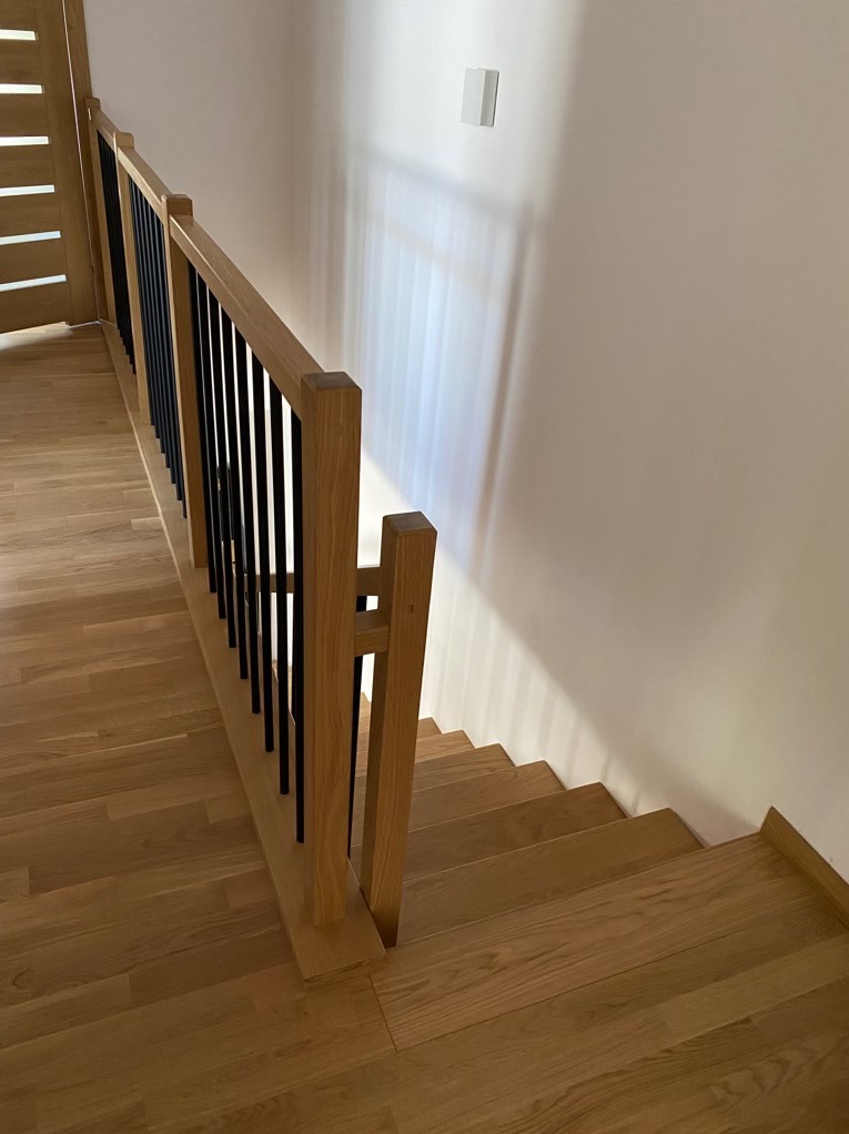 Drewniane schody z balustradą z jasnego drewna i czarnymi metalowymi prętami, widok z góry na dół, subtelne oświetlenie podkreśla strukturę drewna.
