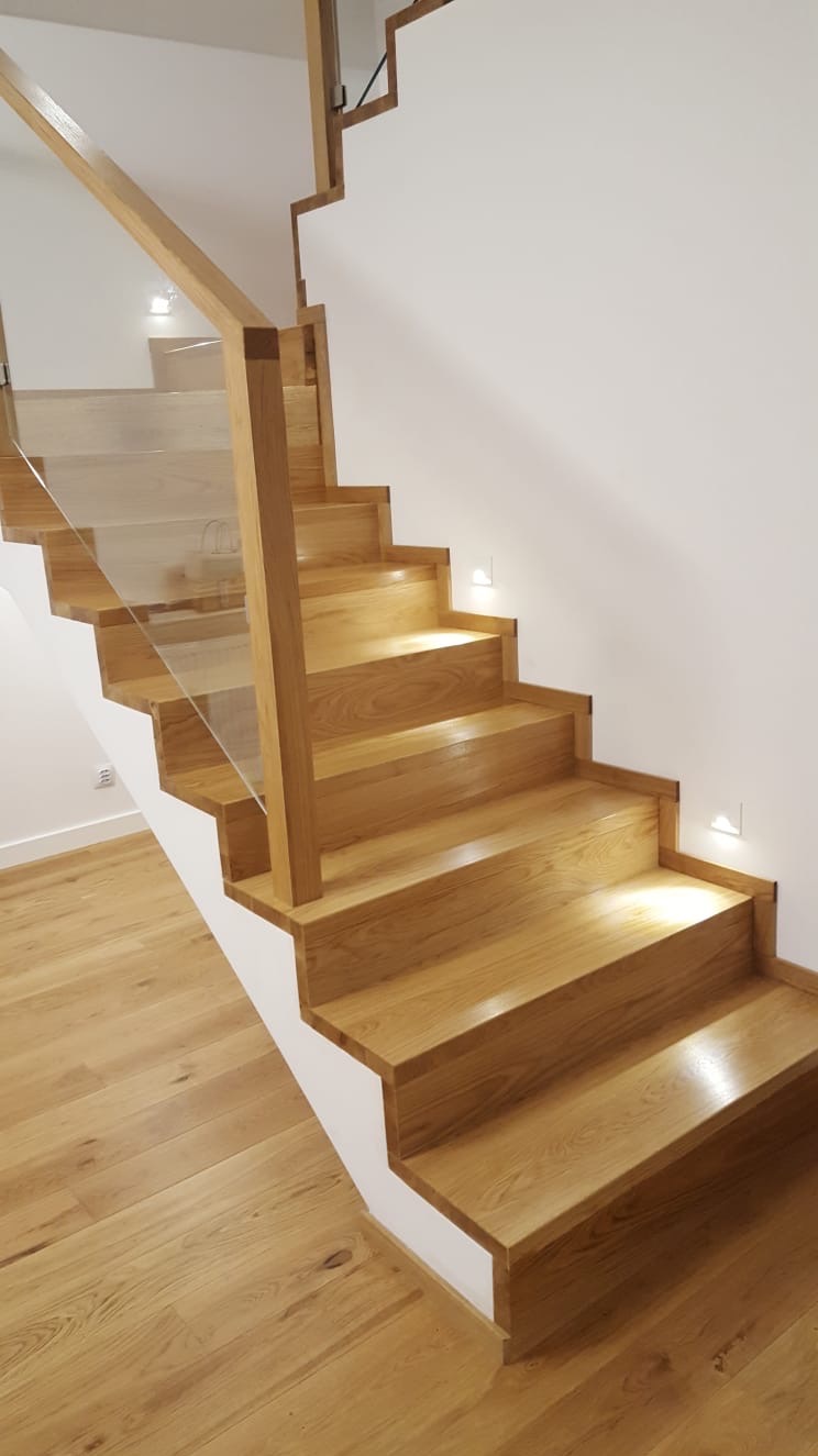 Nowoczesne schody z jasnego drewna z przeszkloną balustradą i podświetleniem LED w ścianie.