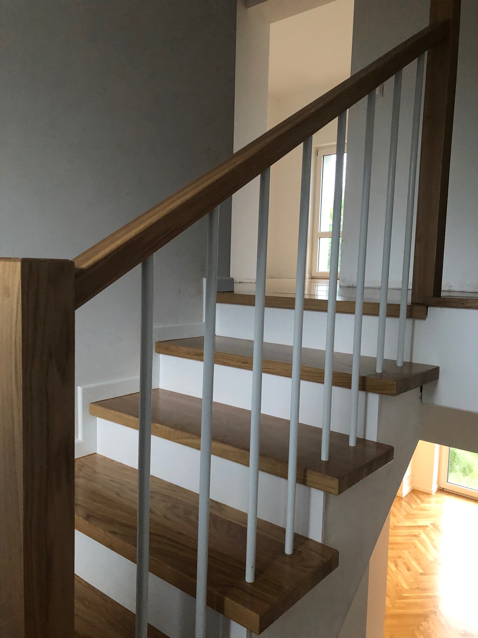 Bieg schodów z drewnianymi stopniami, białymi podstopnicami i balustradą z drewnianą poręczą oraz białymi, pionowymi słupkami, widok z dołu na górę.