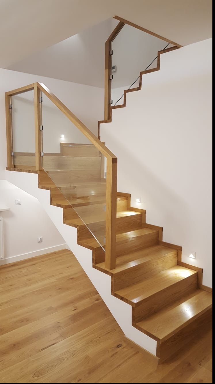 Jasne, drewniane schody z przeszkloną balustradą i punktowym oświetleniem na ścianie, z widocznym fragmentem podłogi wyłożonej parkietem.