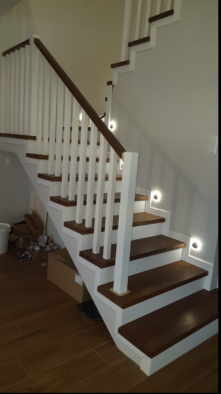 Białe schody z drewnianymi stopniami, oświetlone punktowymi lampkami, z białą balustradą i brązową poręczą. Widok z dołu, na podłodze kartony i śmieci.
