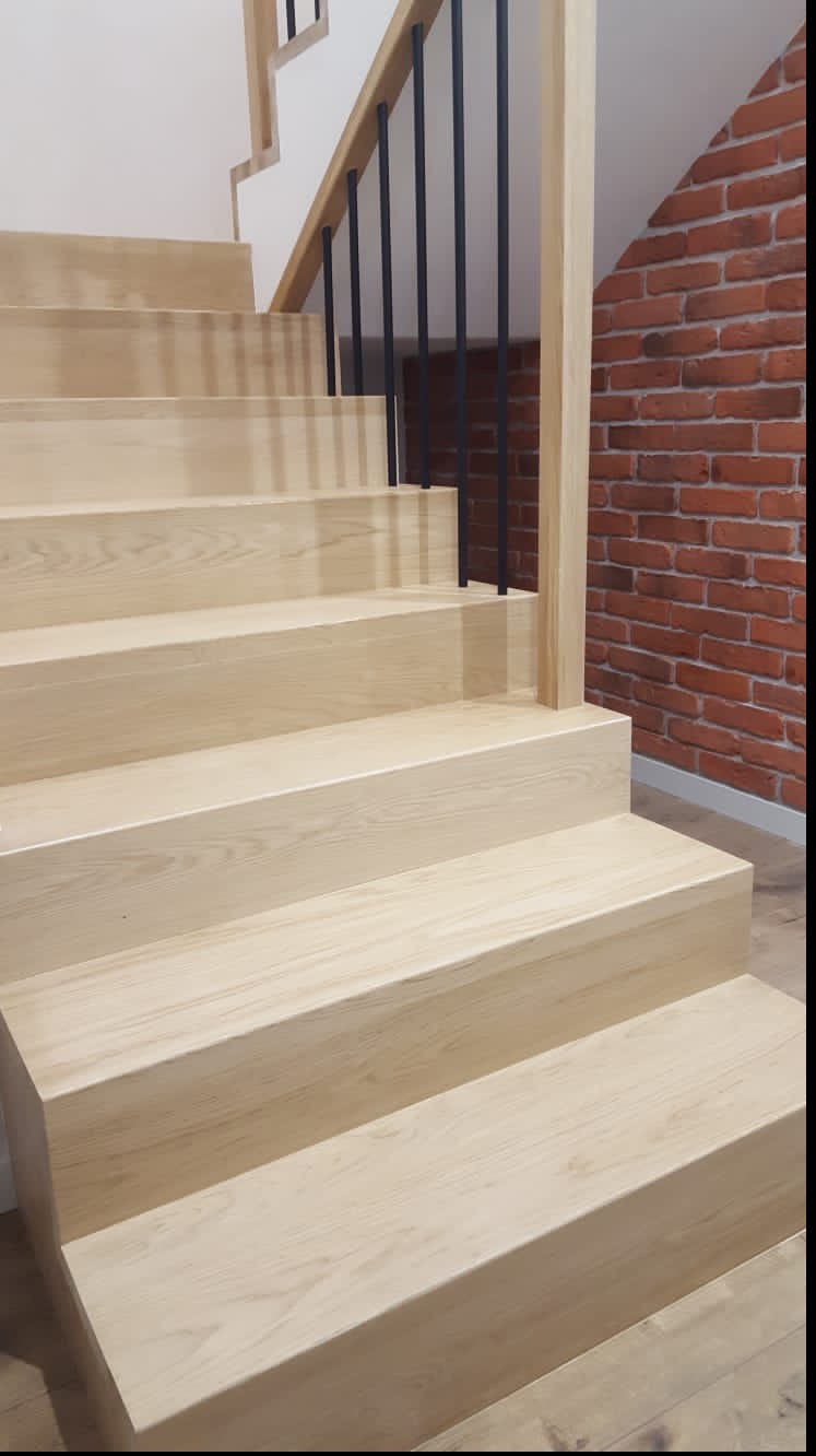 Jasne, drewniane schody z czarnymi, metalowymi słupkami balustrady na tle ściany z czerwonej cegły. Ujęcie od dołu, ukazujące strukturę drewna.