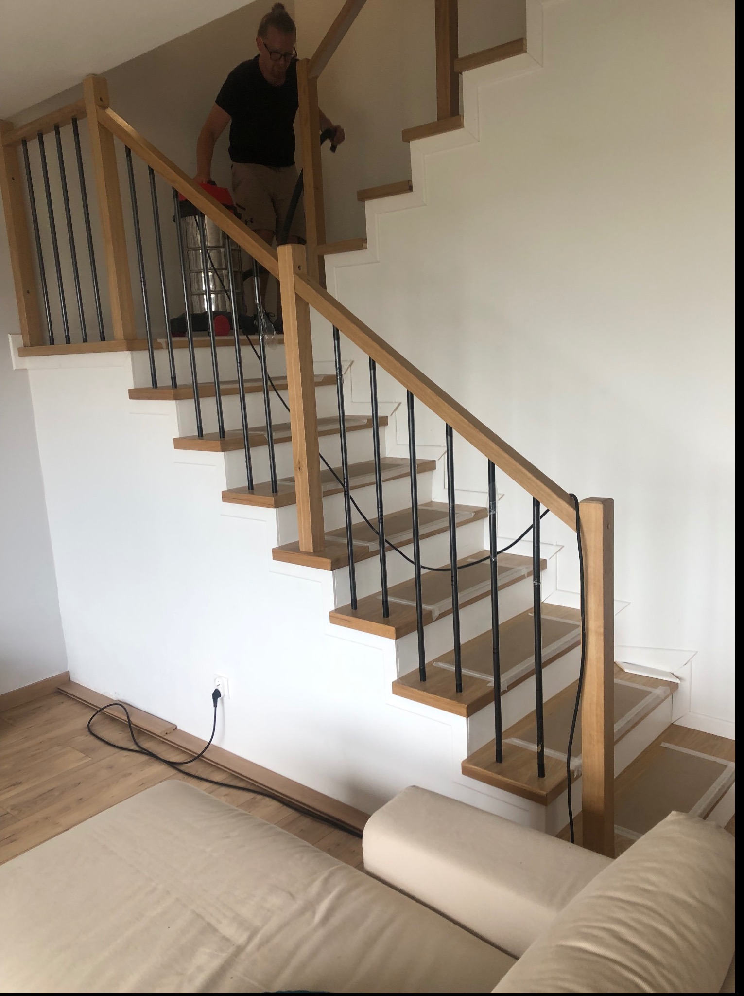 Montaż schodów z drewnianymi stopniami i metalowymi balustradami. Widoczny pracownik z odkurzaczem na schodach. Jasne wnętrze, minimalistyczny design.