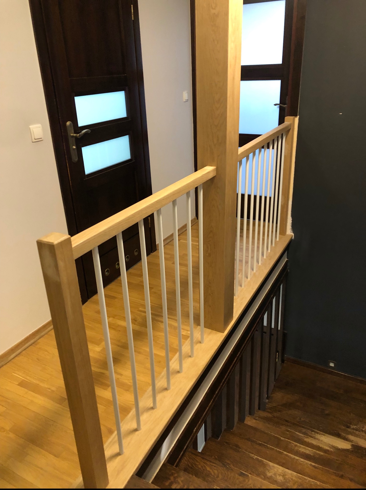 Jasne drewniane schody z białą balustradą w nowoczesnym wnętrzu. Widok z góry na drewniane stopnie i fragment podłogi. Ciemne drzwi w tle.