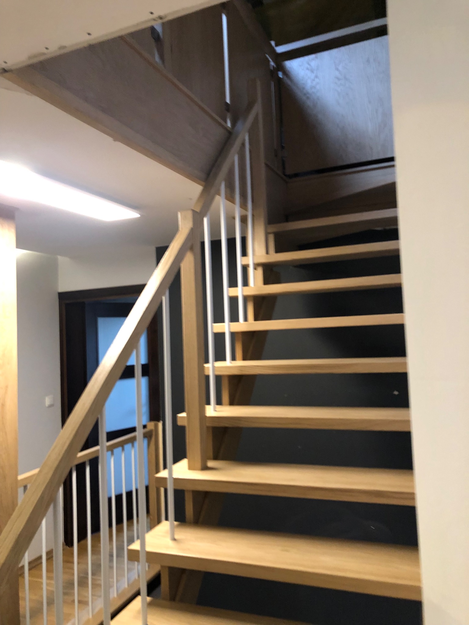 Jasne schody z drewnianymi stopniami i białymi, pionowymi balustradami, prowadzące do otworu w suficie. Widok od dołu, z fragmentem drzwi w tle.
