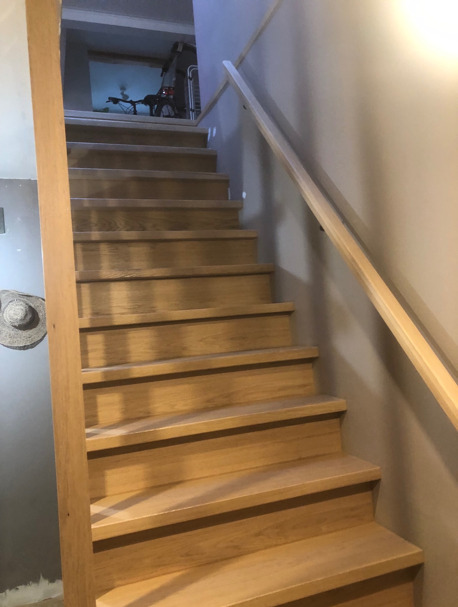 Jasne, drewniane schody z prostą, drewnianą poręczą. Na ścianie wisi słomkowy kapelusz. Na górze schodów widoczny rower.