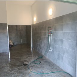 VIVOBUD - Świeżo wykafelkowana łazienka z szarą glazurą, widoczny wąż ogrodowy i instalacja wodna, mokra podłoga z odpływem.