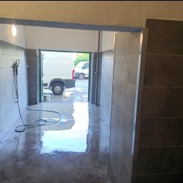 VIVOBUD - Wnętrze myjni samochodowej z szarymi płytkami na ścianach, mokra podłoga i wąż ciśnieniowy zwisający ze ściany, widoczne białe busy w tle przez otwarte wrota.