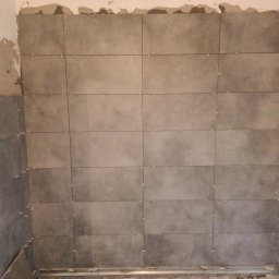 VIVOBUD - Ściana w trakcie układania szarych płytek ceramicznych, widoczne krzyżyki dystansowe i fragment surowej ściany z resztkami zaprawy.