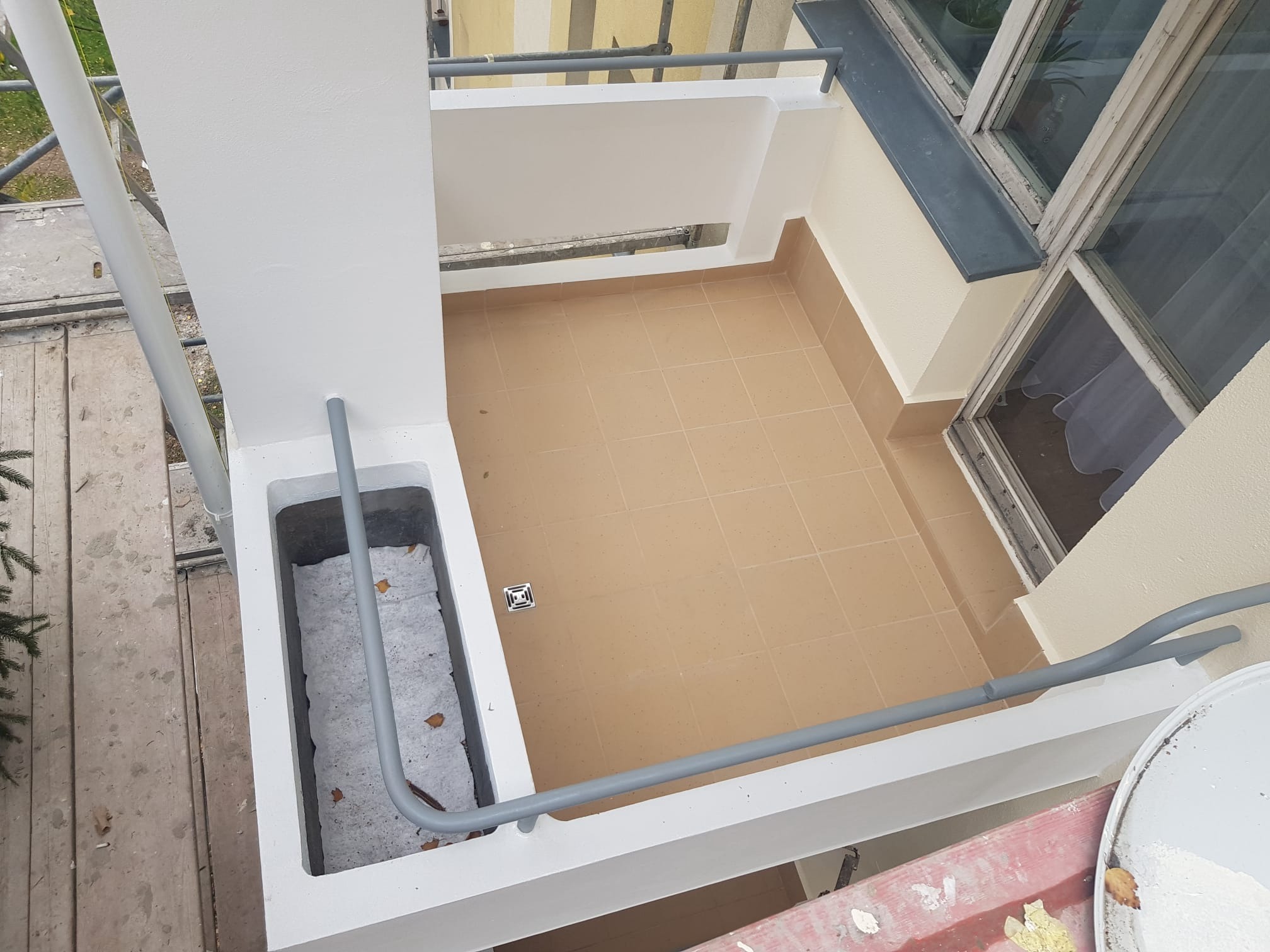 Pomalowany na biało balkon z brązowymi płytkami podłogowymi, widoczne rury odpływowe i kratka ściekowa, fragment rusztowania po lewej stronie.