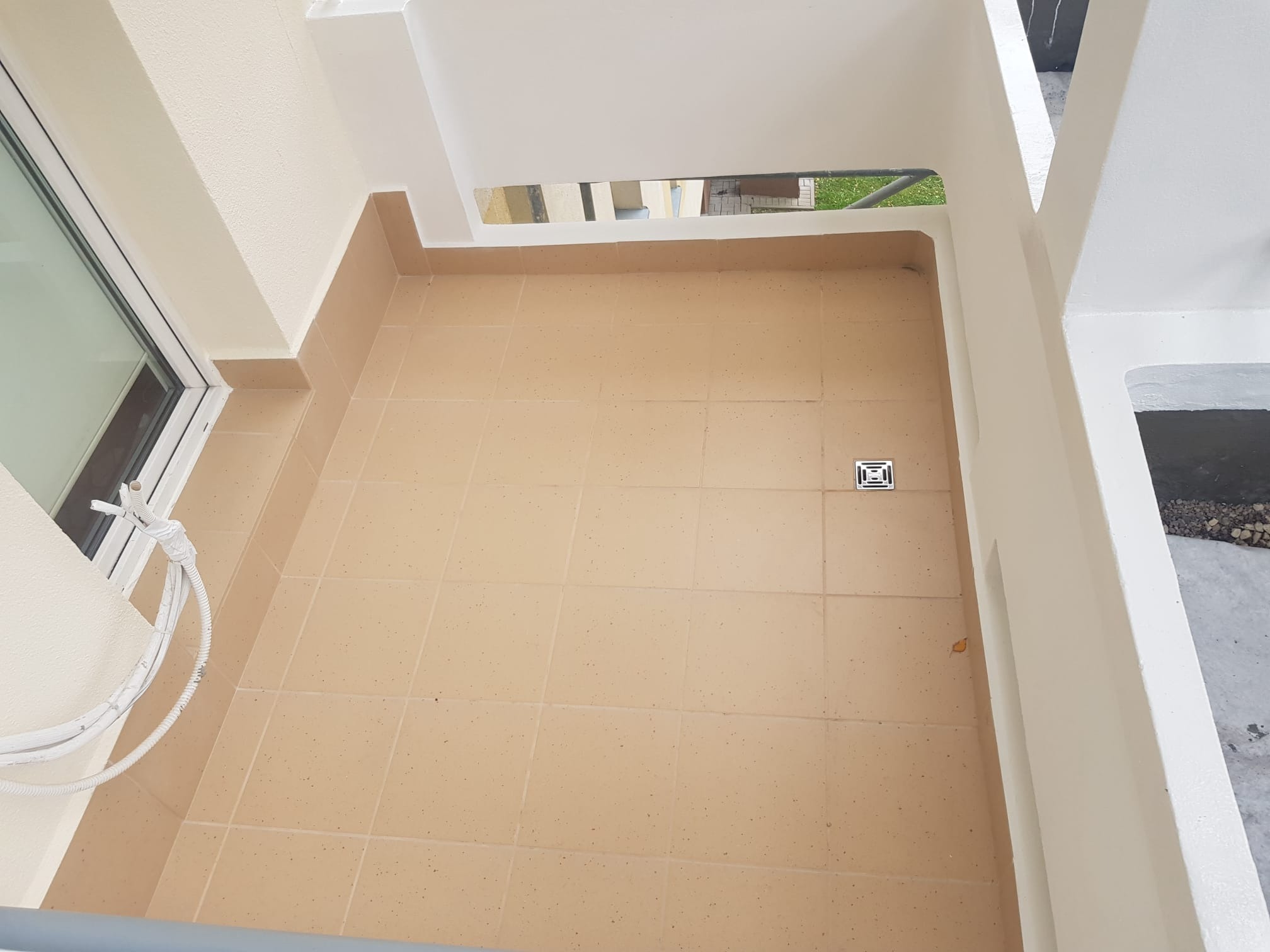 Wykafelkowany balkon z beżowymi płytkami, widok z góry, białe ściany i odpływ liniowy w podłodze, częściowo widoczne okno i przewody elektryczne.