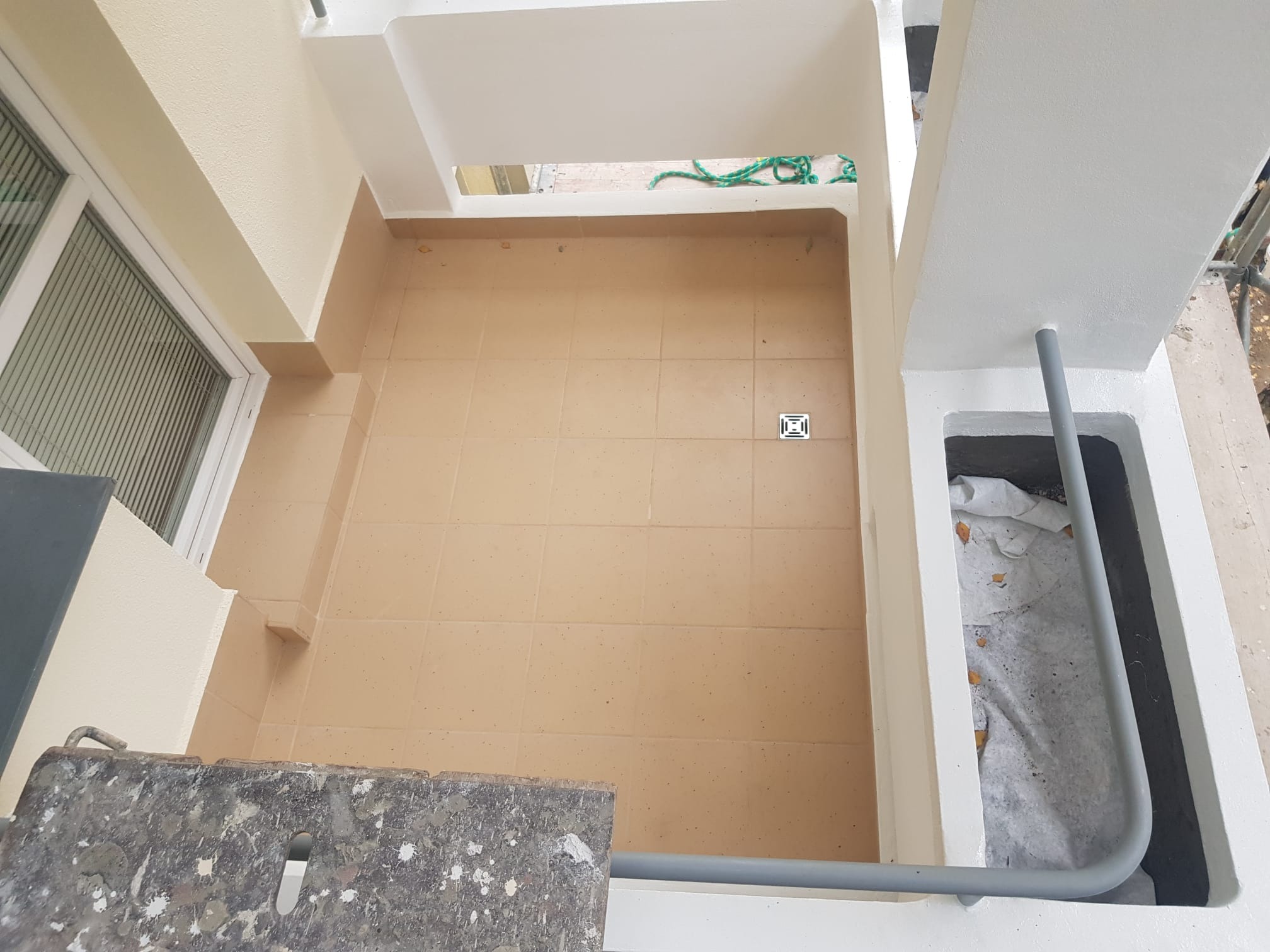 Widok z góry na wykafelkowany balkon po remoncie, z widoczną kratką odpływową, świeżo pomalowanymi ścianami i fragmentem drabiny malarskiej.