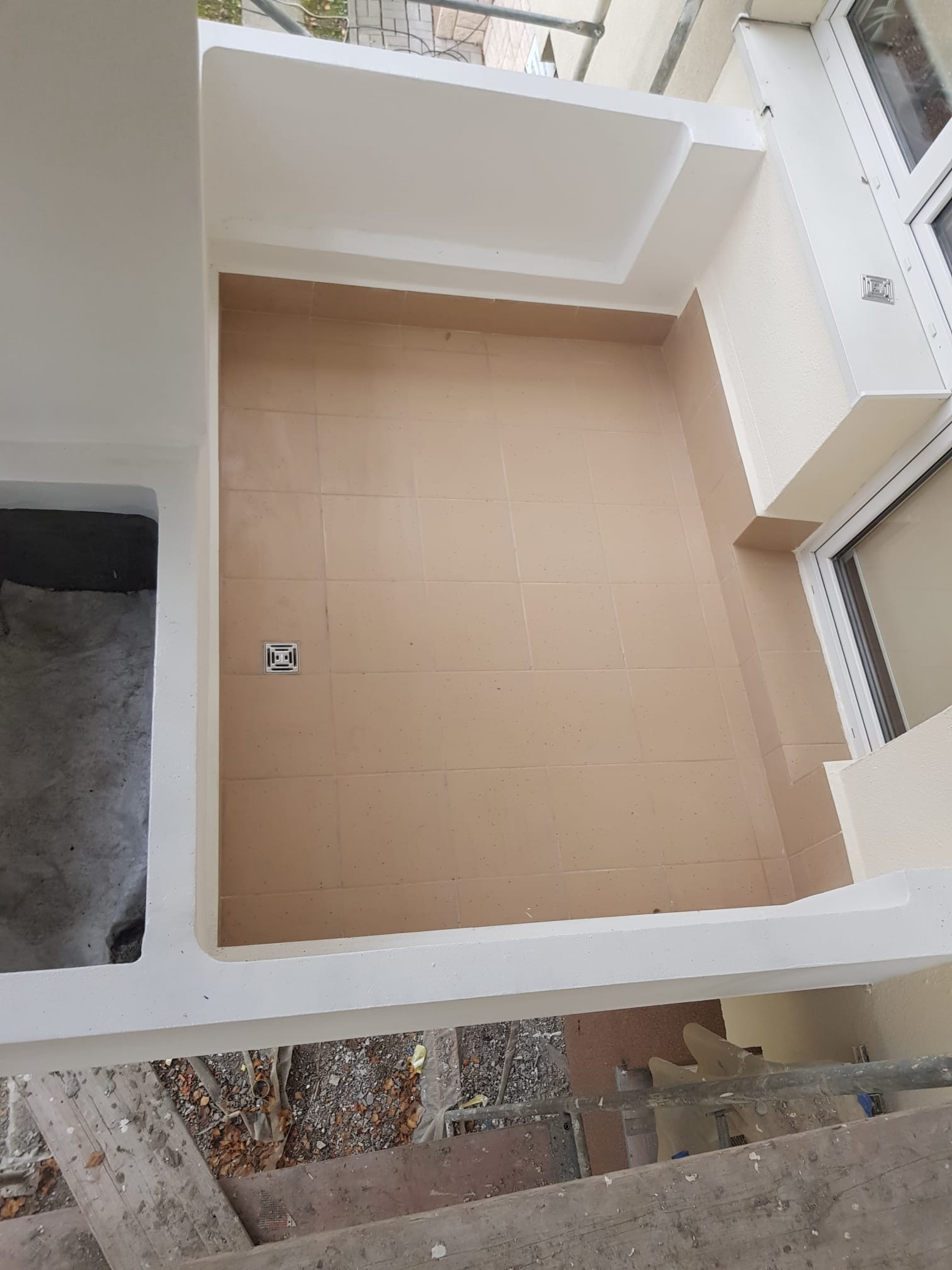 Wykończony brodzik z beżowymi płytkami i odpływem liniowym, białe ściany i rama okienna, widok z góry z rusztowania.