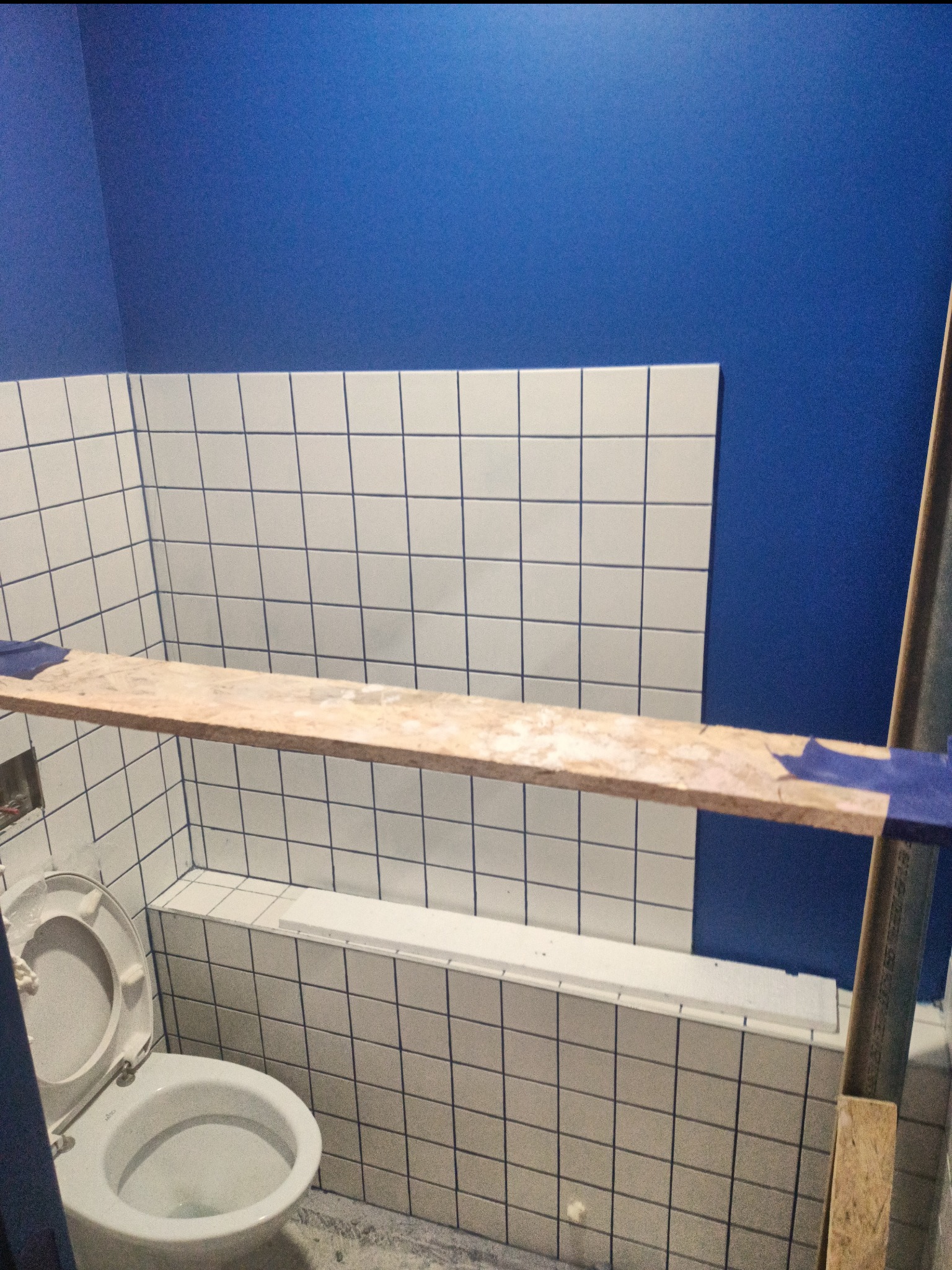Błękitna ściana nad białą, kwadratową glazurą z ciemnoniebieską fugą w trakcie prac wykończeniowych w łazience, widoczny fragment toalety i drewniana deska na rusztowaniu.