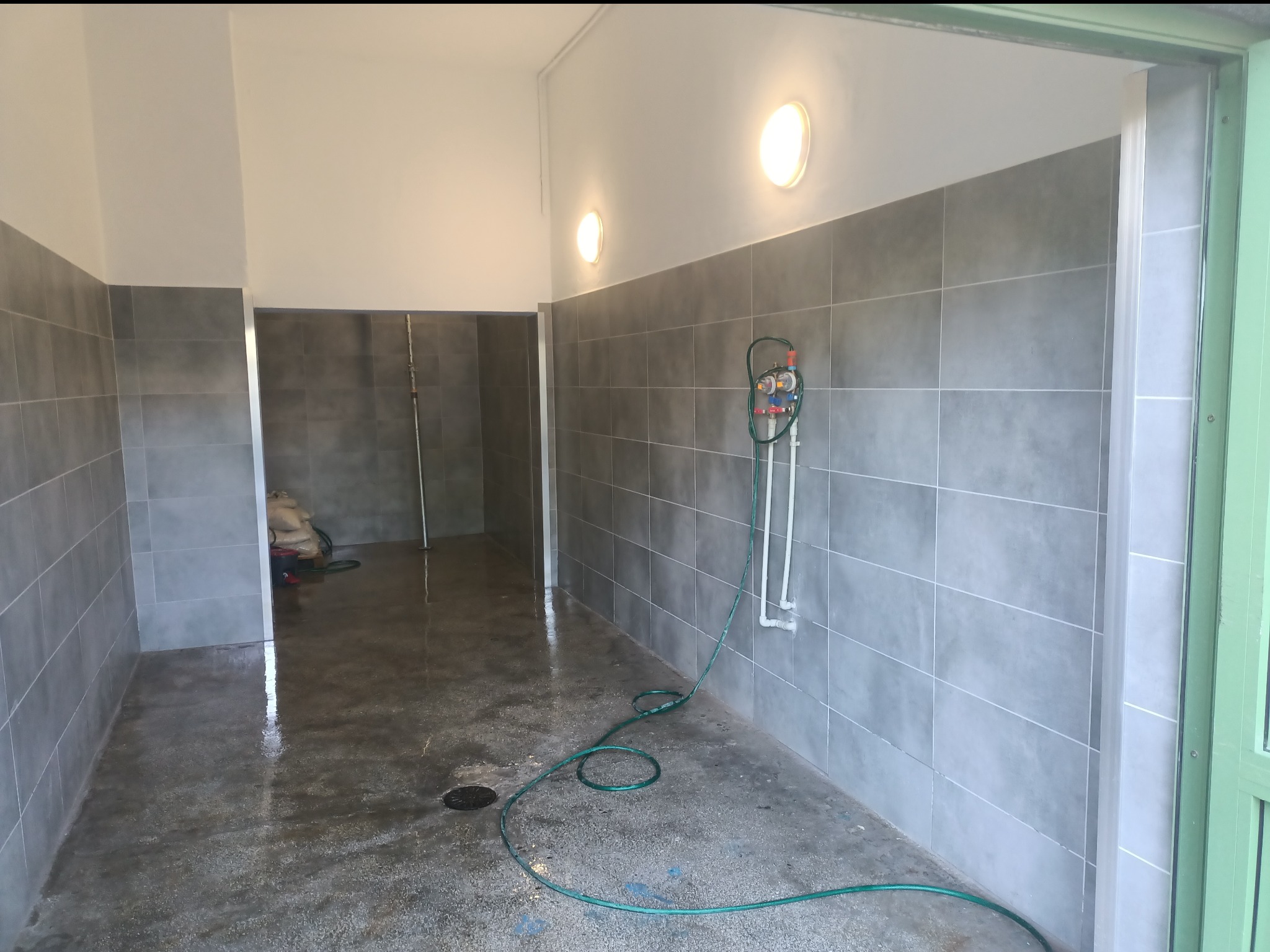 Świeżo wykafelkowana łazienka z szarą glazurą, widoczny wąż ogrodowy i instalacja wodna, mokra podłoga z odpływem.