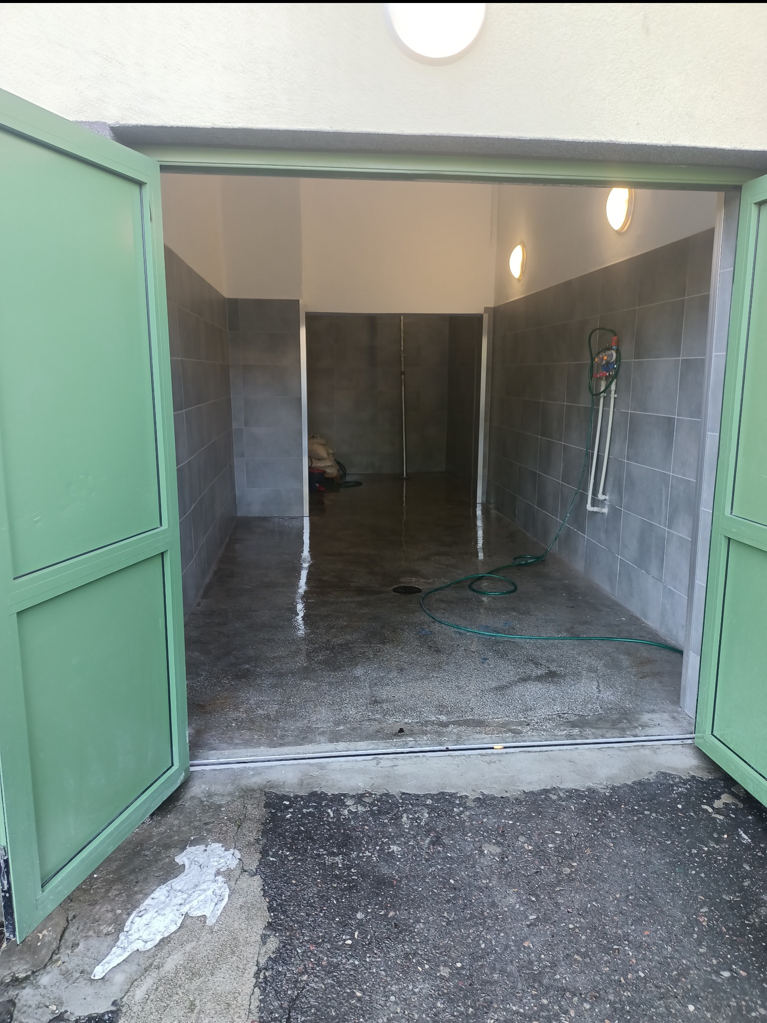 Wnętrze umytego pomieszczenia z szarymi płytkami, widoczne otwarte, zielone drzwi i mokra posadzka, na zewnątrz plama po cemencie.