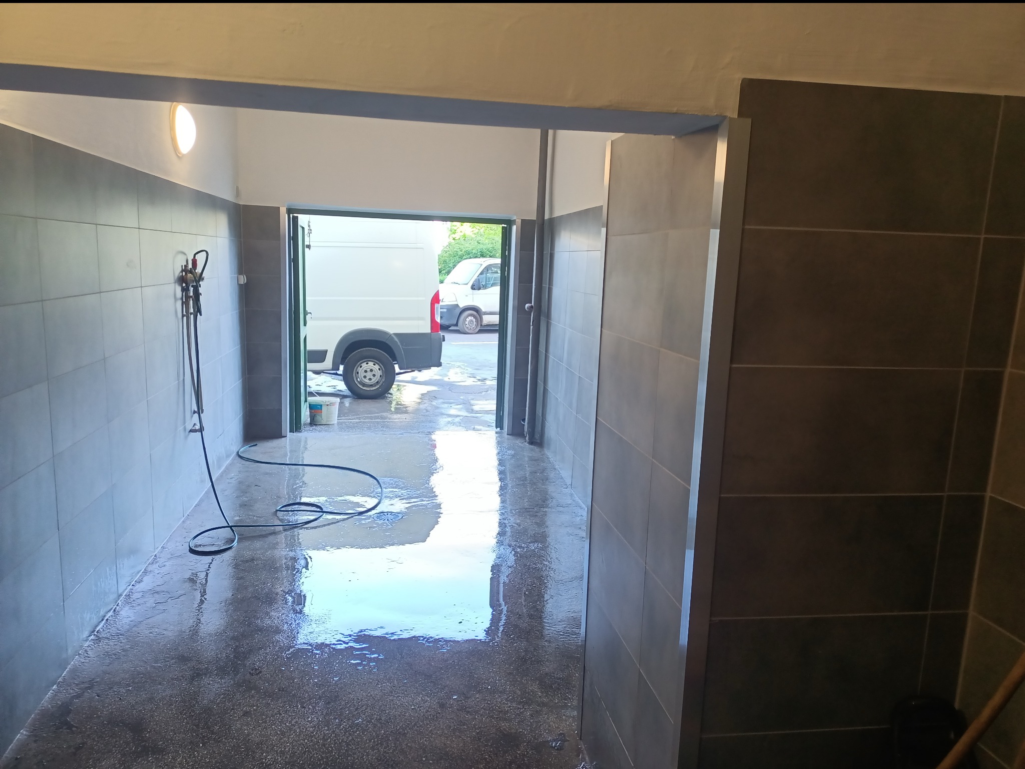 Wnętrze myjni samochodowej z szarymi płytkami na ścianach, mokra podłoga i wąż ciśnieniowy zwisający ze ściany, widoczne białe busy w tle przez otwarte wrota.