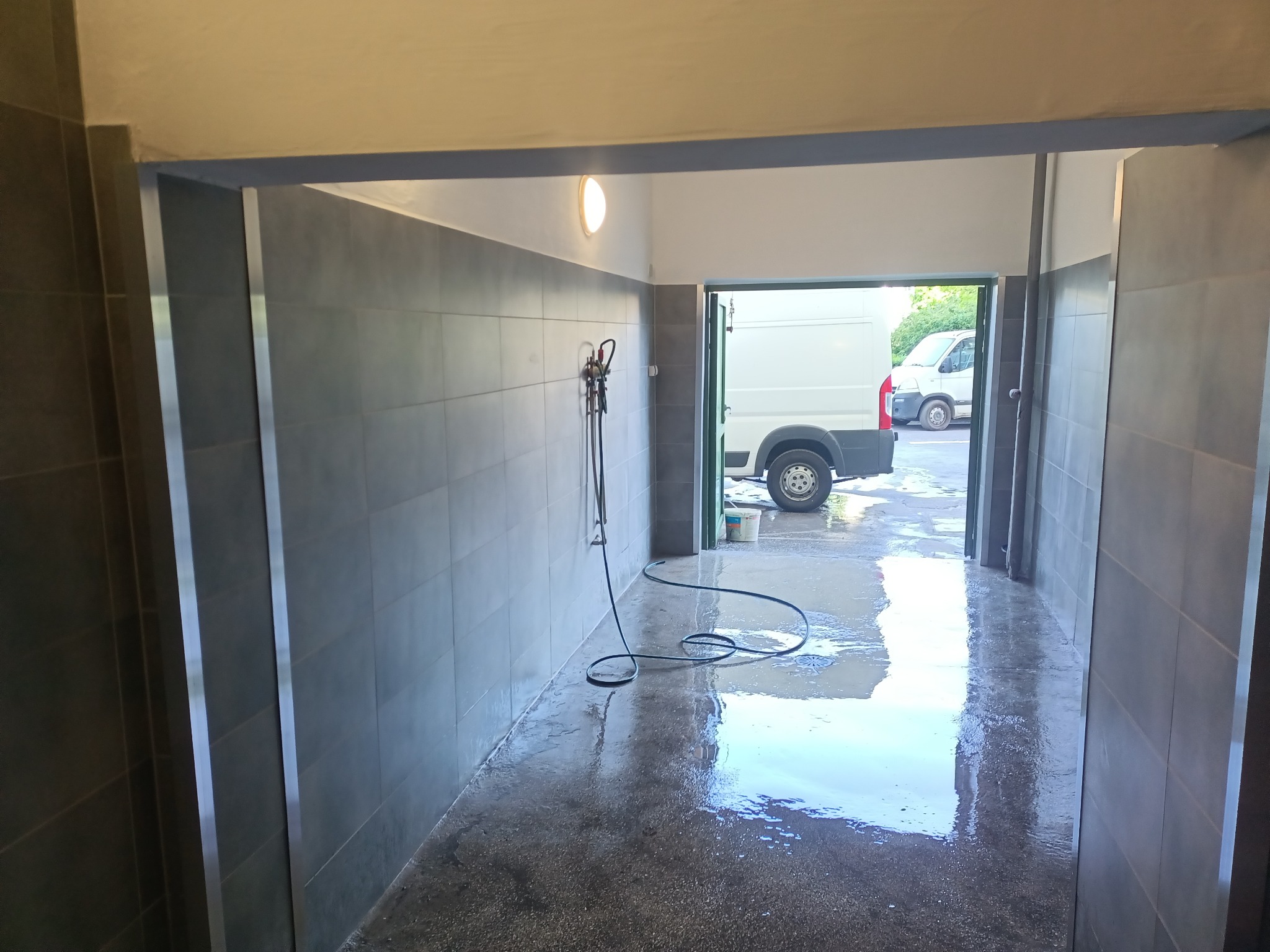 Wnętrze myjni samochodowej z szarymi płytkami, mokrą podłogą i wężem natryskowym, widok na biały samochód dostawczy na zewnątrz.