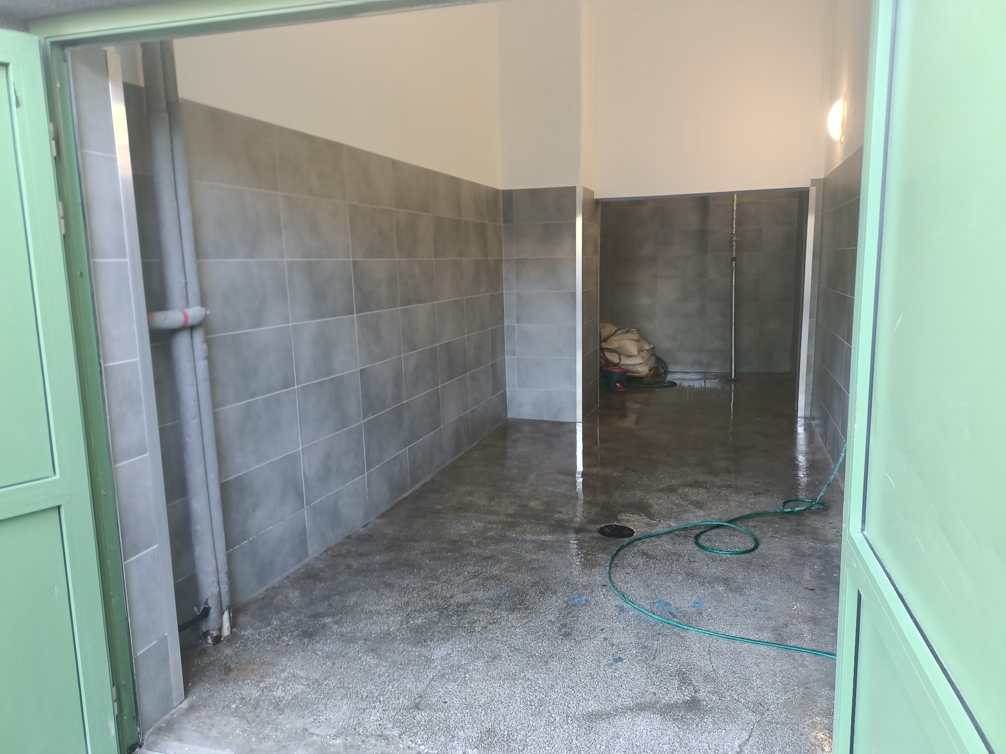 Świeżo wykafelkowane pomieszczenie łazienkowe w szarościach, widok z zewnątrz przez otwarte, zielone drzwi, mokra posadzka z odpływem, w tle worki z materiałami budowlanymi.